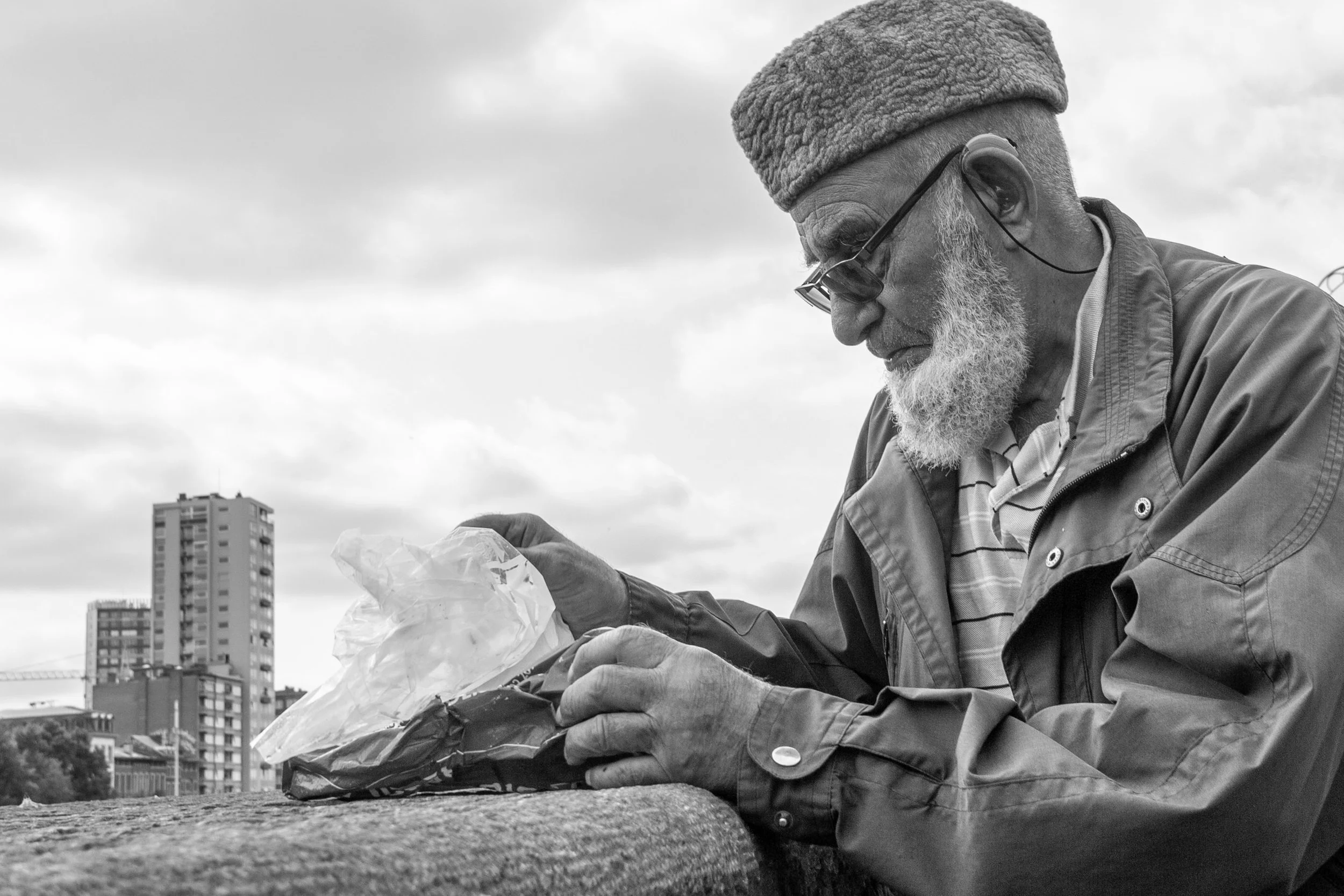 An elderly man with a beard and glasses, wearing a warm hat and jacket, is looking at and holding a plastic bag while sitting outdoors with a cityscape of tall buildings in the background.