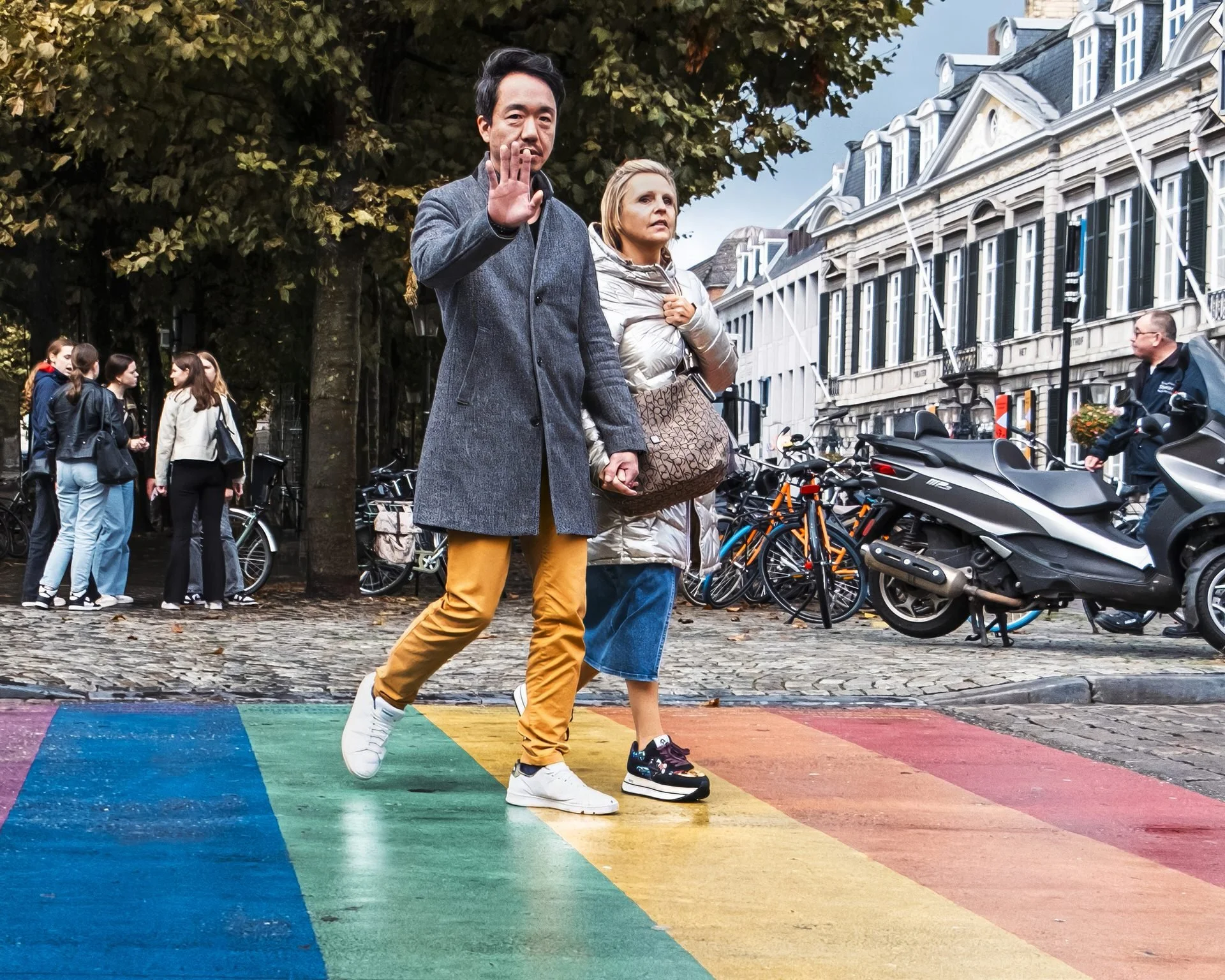 A couple holding hands walking on a rainbow-colored crosswalk in an urban area, with several people, bicycles, and parked motorcycles nearby, and historic buildings in the background.