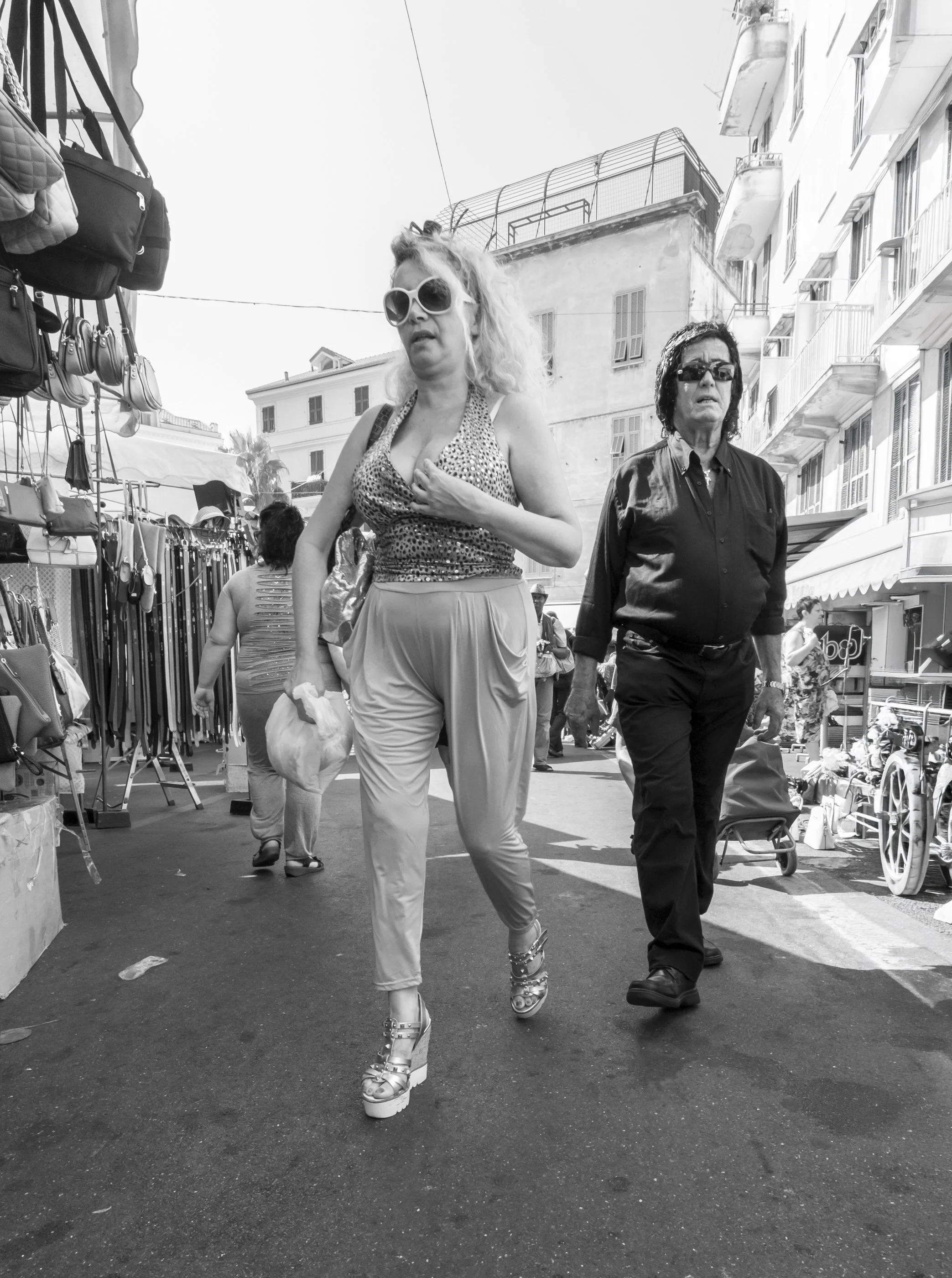 Two women walking through a street market; one wearing large sunglasses, high heels, and a leopard print top, the other in dark sunglasses and a black shirt; market stalls and buildings in the background.