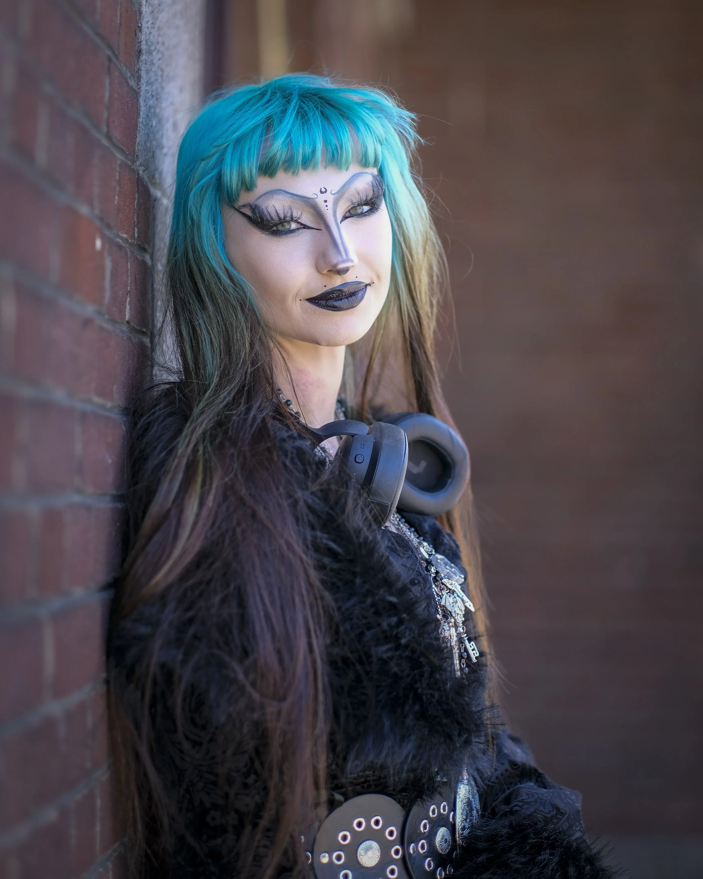 A woman with striking blue hair and elaborate black makeup on her face, leaning against a brick wall. She has a detailed facial design with black and white accents, and her eyes are enhanced with long false eyelashes. She is wearing dark clothing and