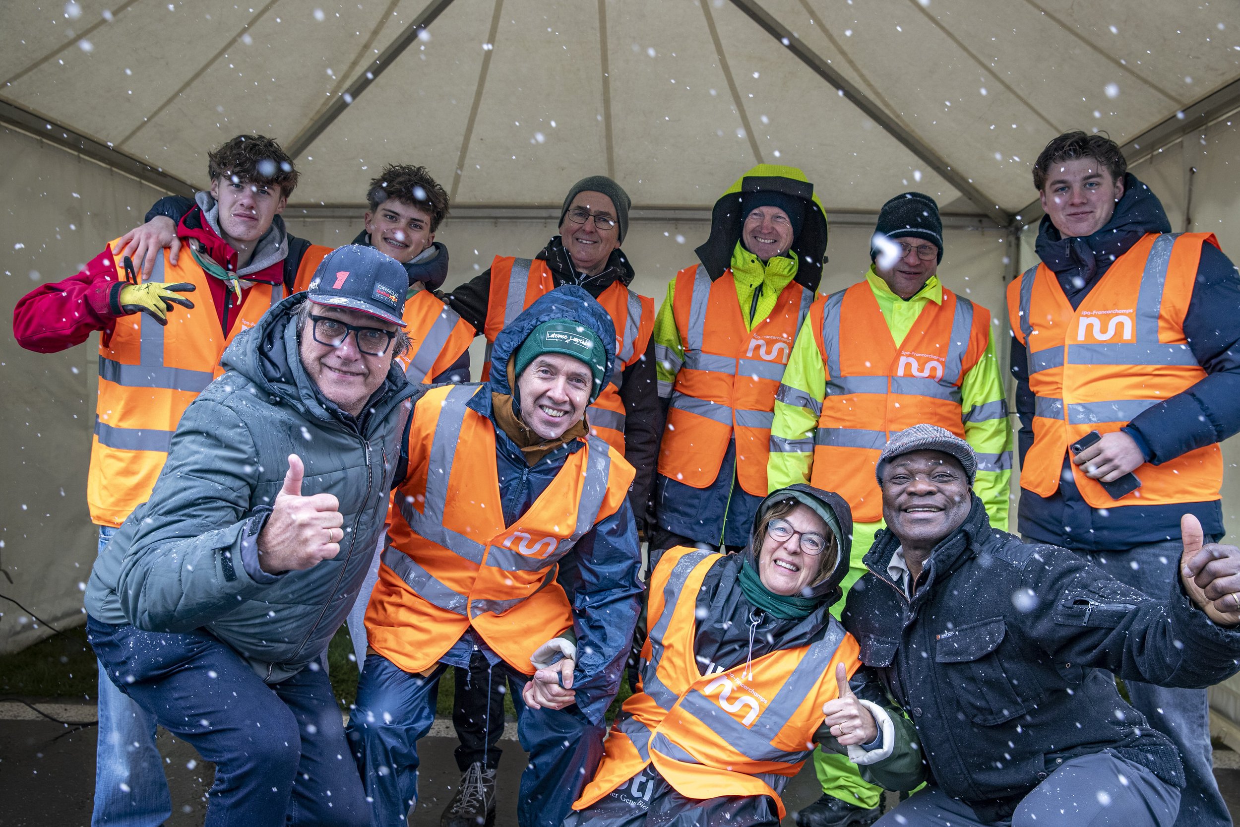 Group of people in orange safety vests and winter clothing posing under a beige tent with snow falling.
