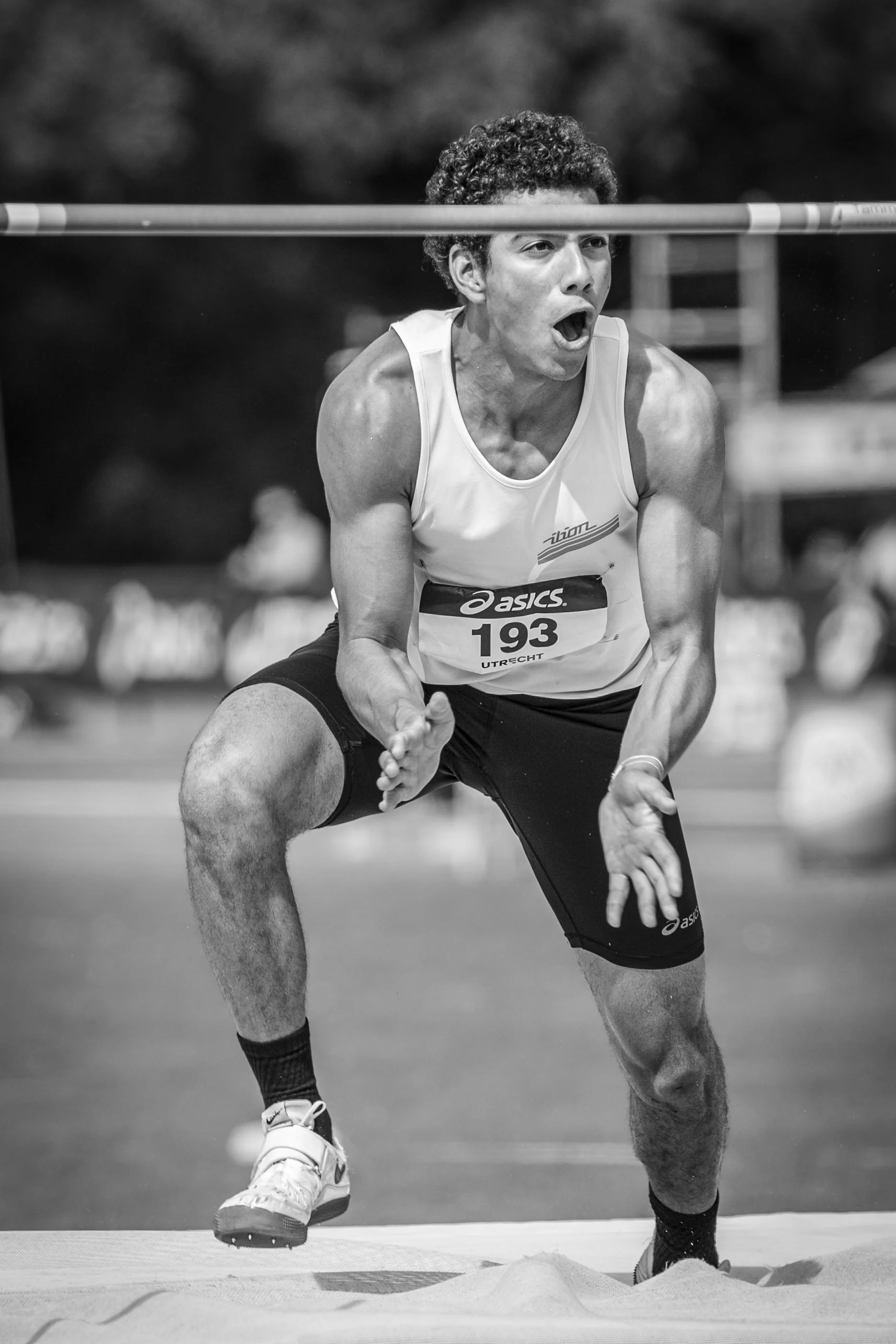 A male athlete with curly hair participating in a long jump event, mid-air, with sand on the ground below.