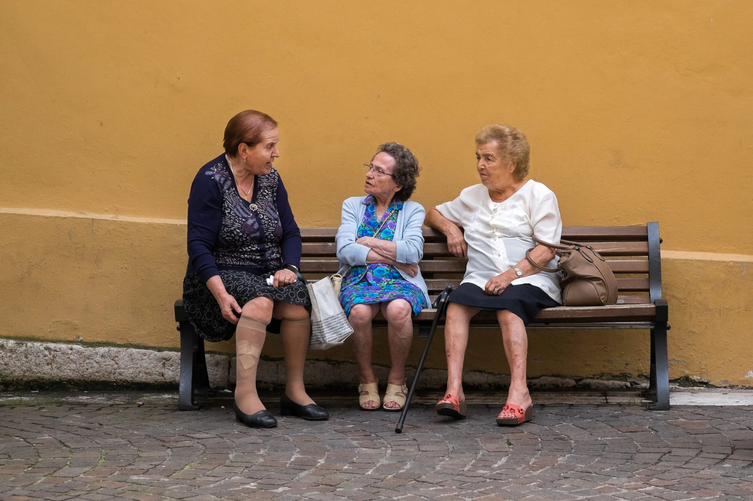 Three elderly women sitting on a wooden bench against a yellow wall having a conversation.