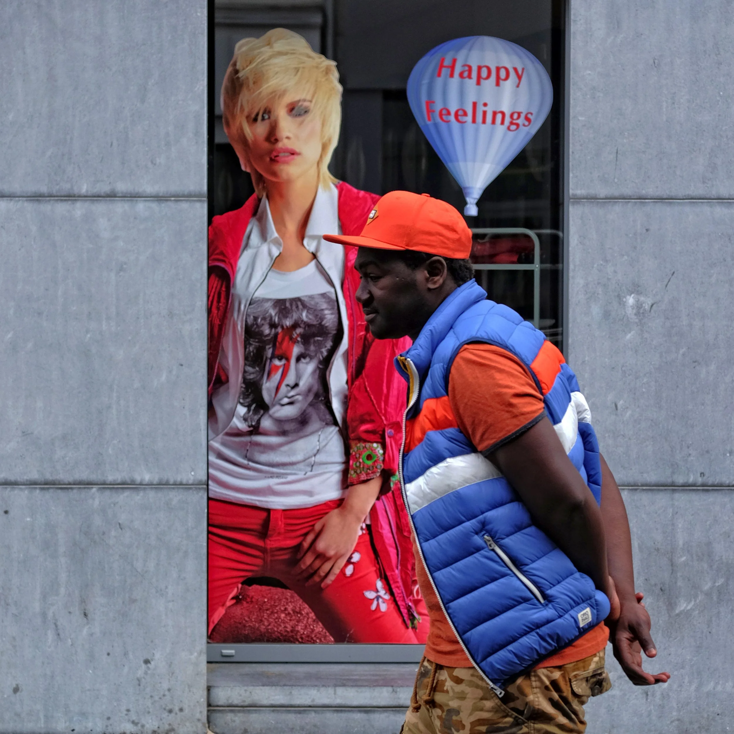 A young woman with blonde hair and a young boy wearing a blue puffer vest and camouflage shorts standing outside near a building with a glass window that has a balloon-shaped sign reading "Happy Feelings" in red letters.