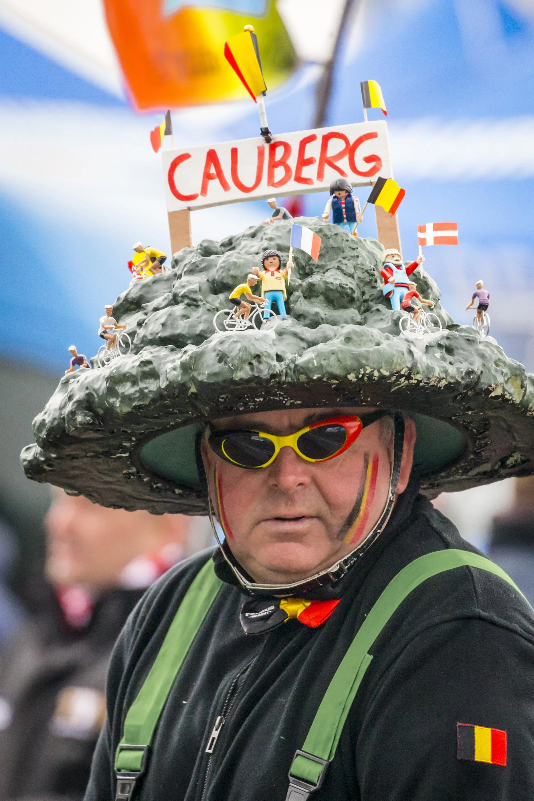 A man wearing cycling gear and a large rock-shaped hat decorated with miniature figures, flags, and a sign that reads 'CĂUBERG'.
