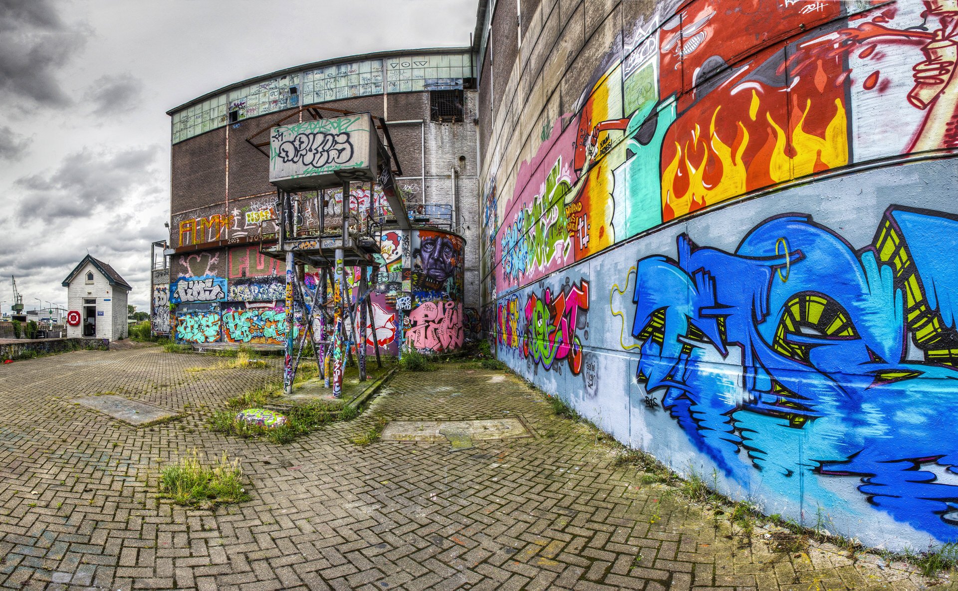 A large brick building covered in colorful graffiti street art, with a cloudy sky overhead. The foreground features a paved area with weeds growing between the tiles.