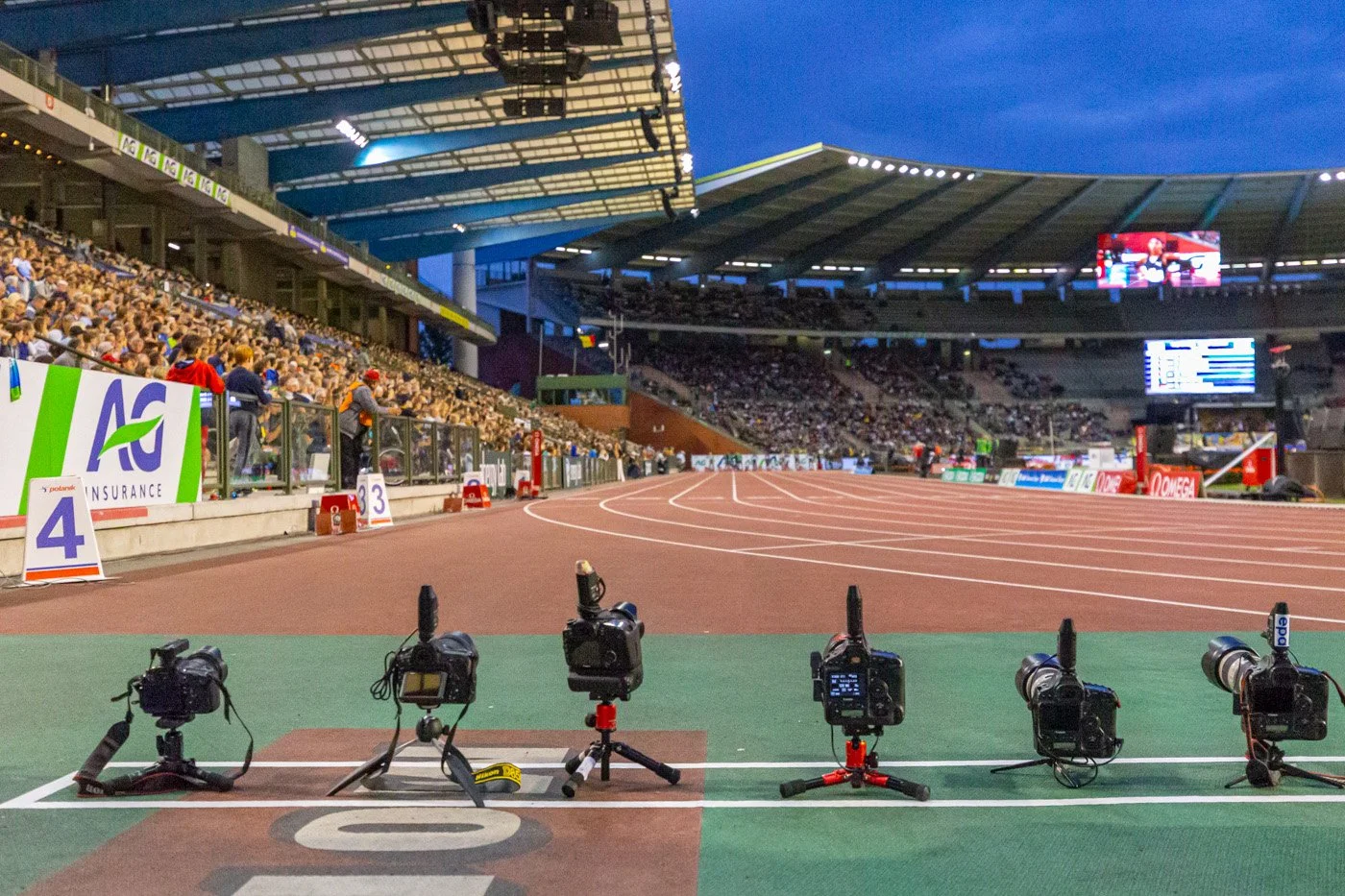 View of a crowded athletics stadium with cameras set up on the track for recording, a large audience seated in the stands, and digital screens displaying event information, during evening or night conditions.