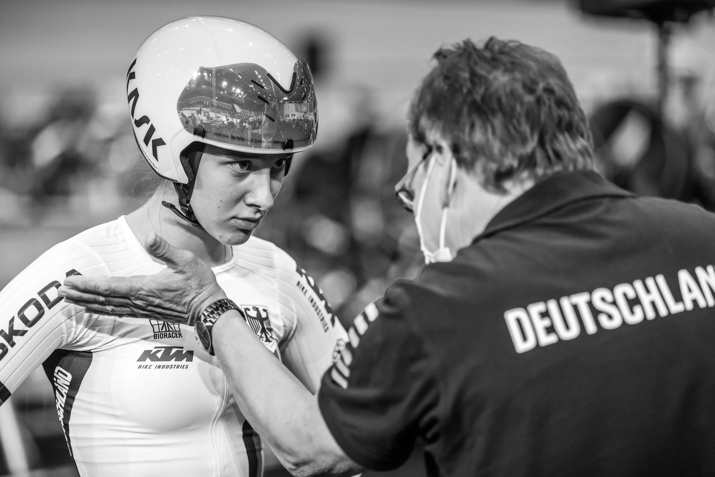 A young female athlete wearing a helmet and sports uniform listening to a man with glasses and face mask, possibly a coach, in what appears to be a sports arena or event setting.