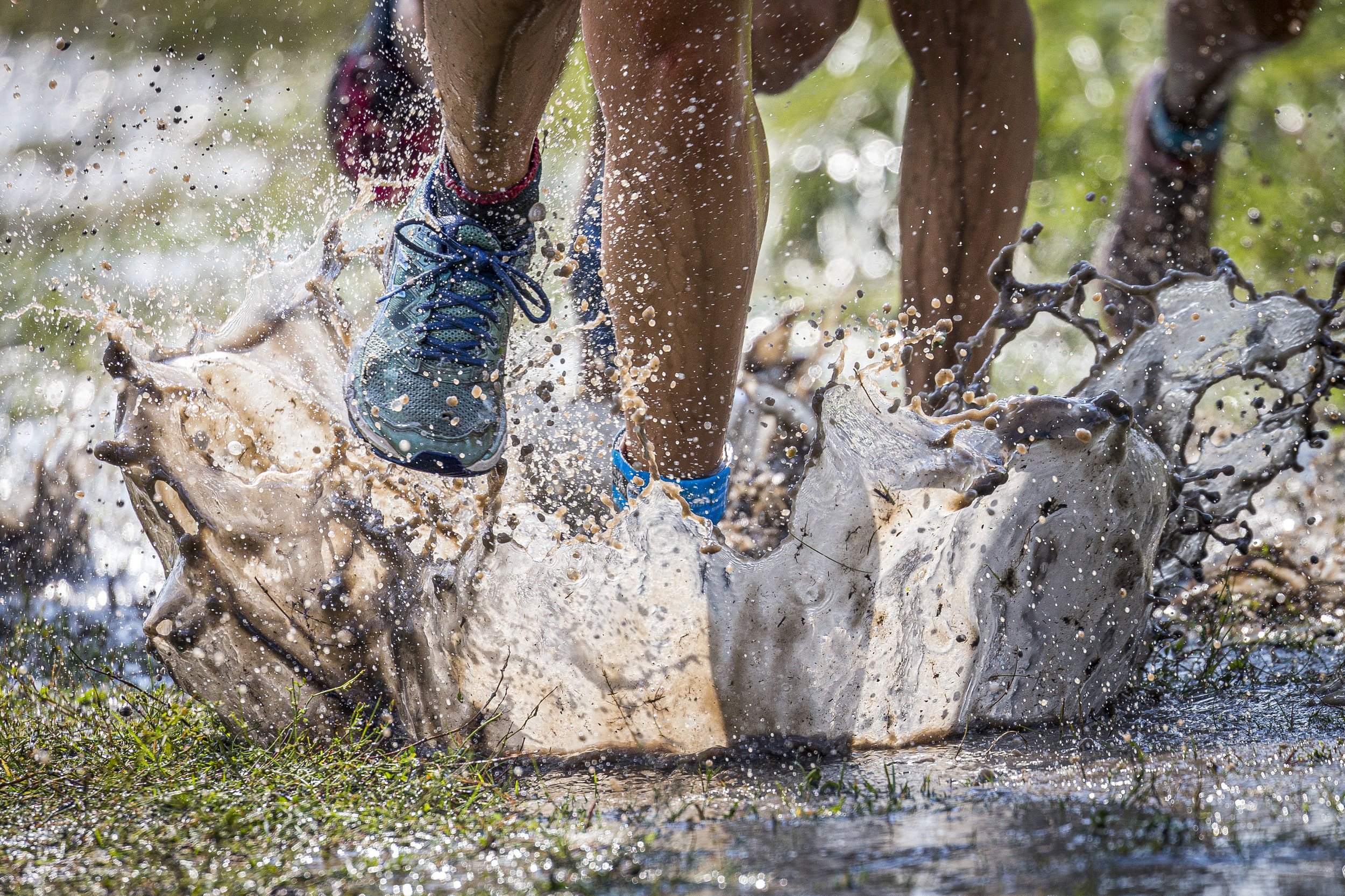 Close-up of a person's legs and feet in blue running shoes splashing through muddy water in a muddy outdoor setting.