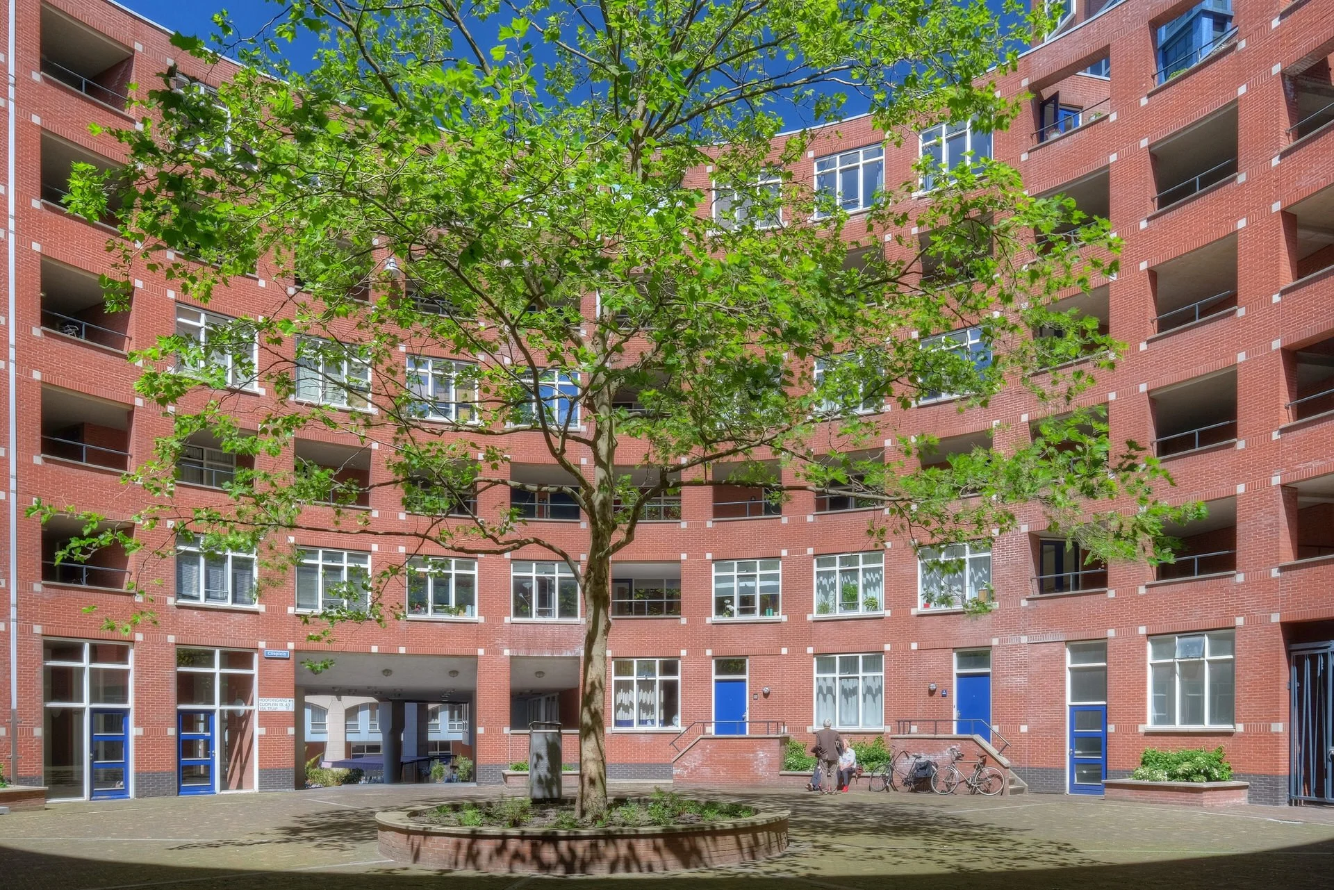 A red brick residential building with multiple balconies and windows, and a small courtyard with a tree, benches, and bicycles in front.