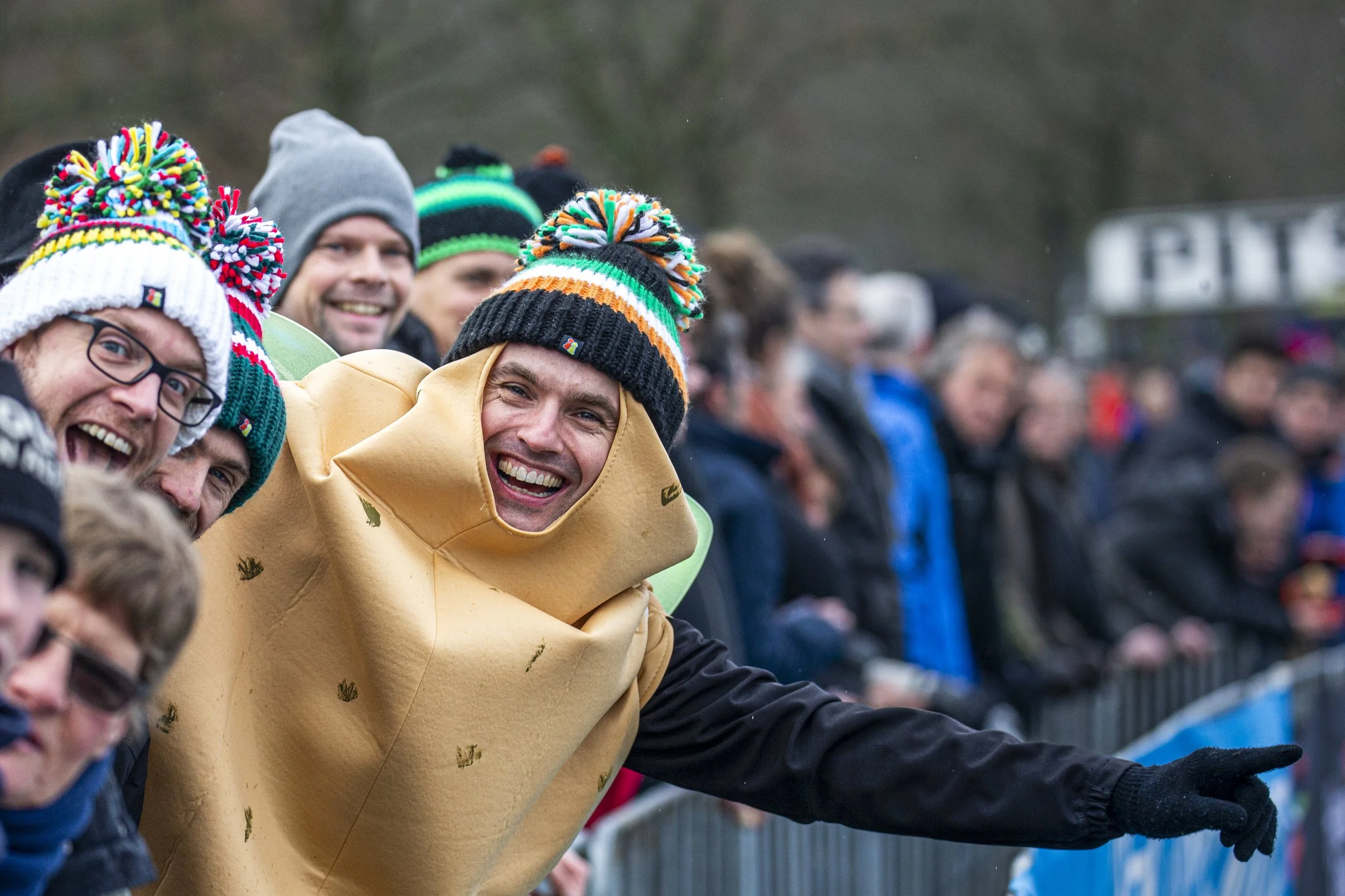 People wearing colorful winter hats and jackets at an outdoor event, smiling and pointing.