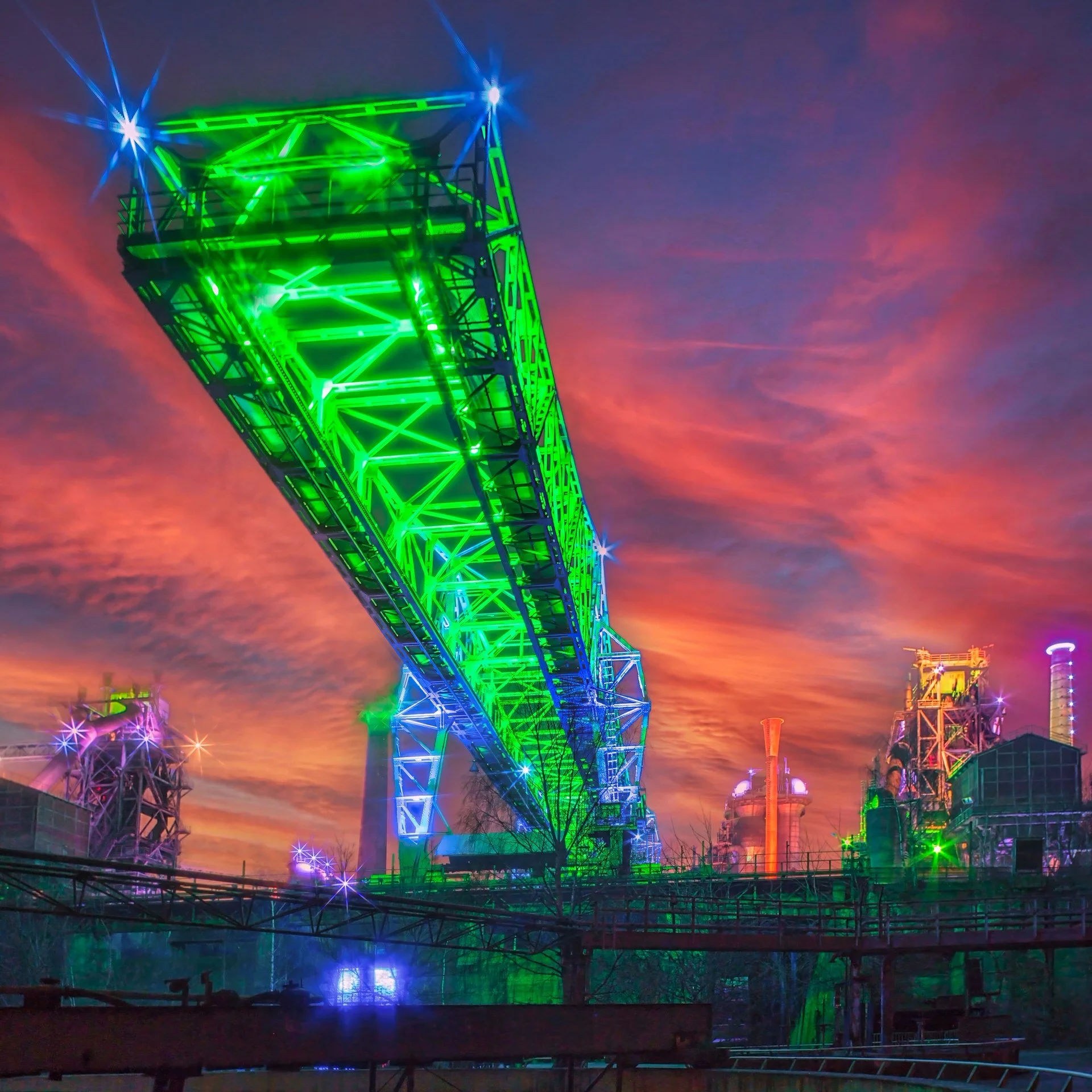 A roller coaster illuminated with bright green, blue, and purple lights against a colorful evening sky with clouds.