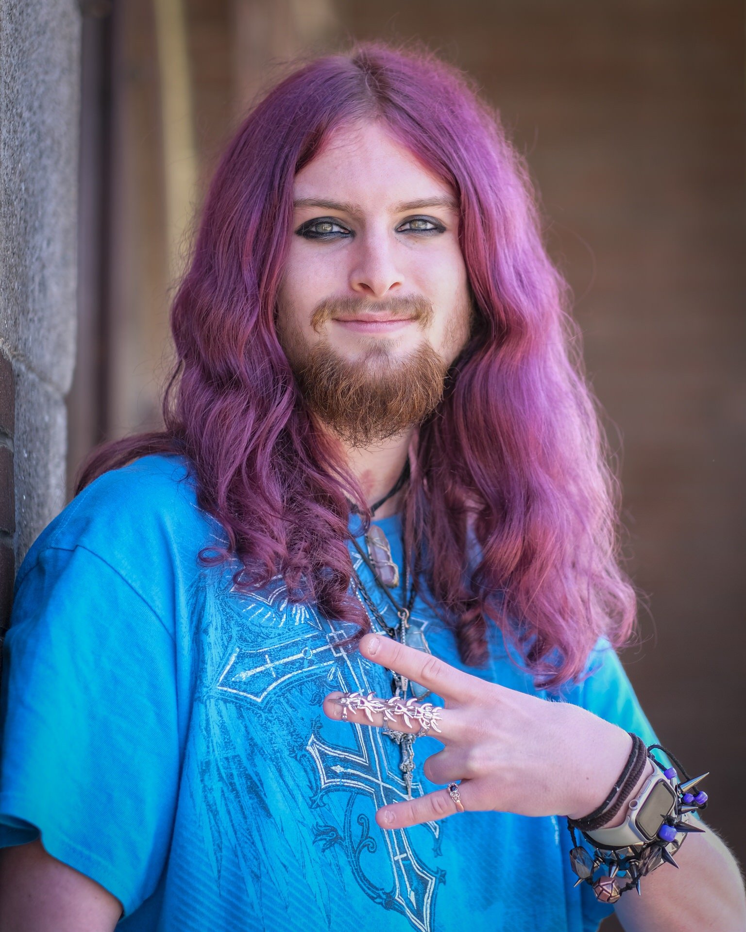 Close-up of a person with long pink-purple hair, a beard, blue eyes, and black eye makeup, wearing a blue shirt and various bracelets, pointing with their index finger.