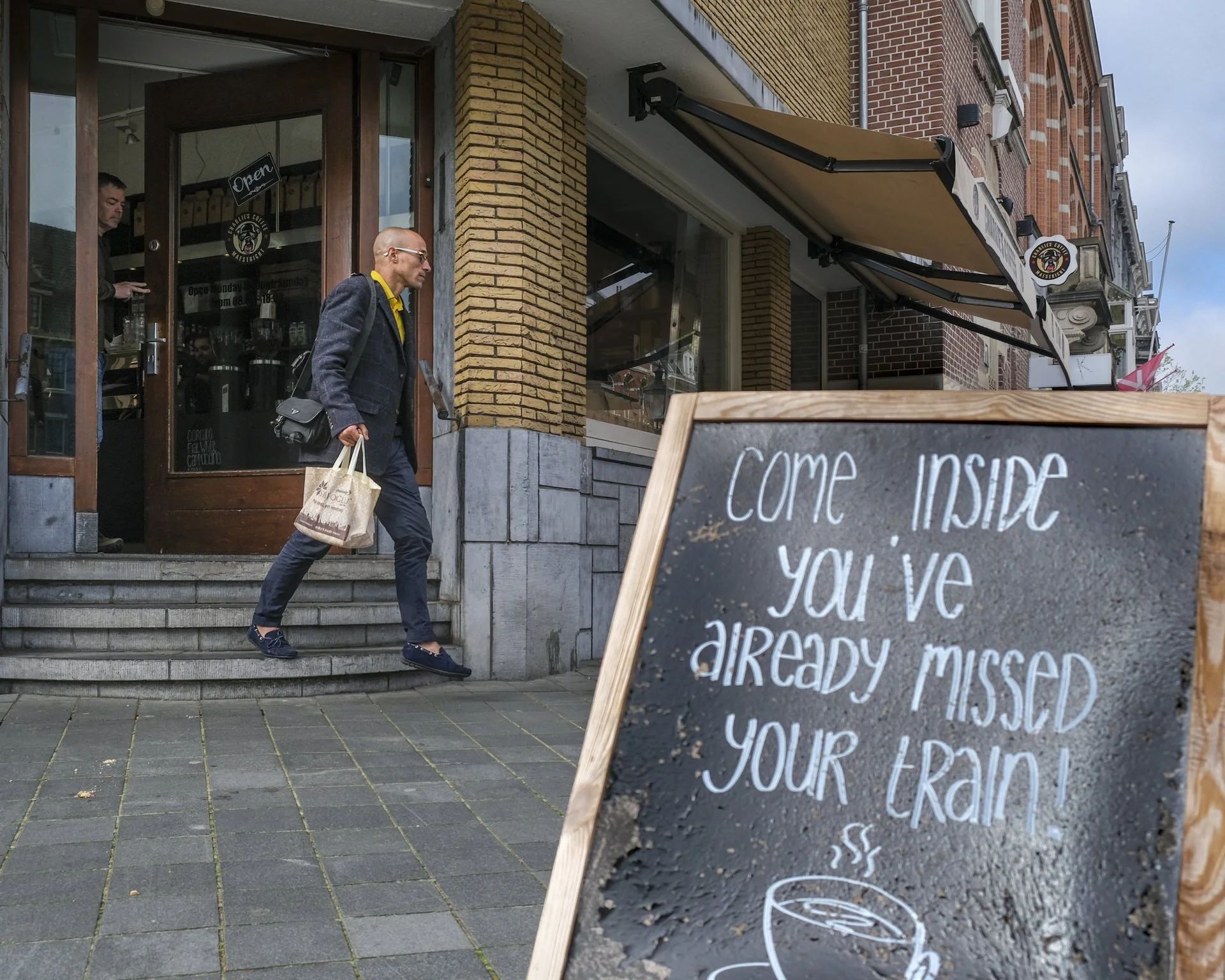 People entering a cafe or restaurant with a chalkboard sign in front that contains a handwritten menu or message. The establishment has a brick exterior with large glass windows and an overhanging awning, and the door is open, welcoming customers ins