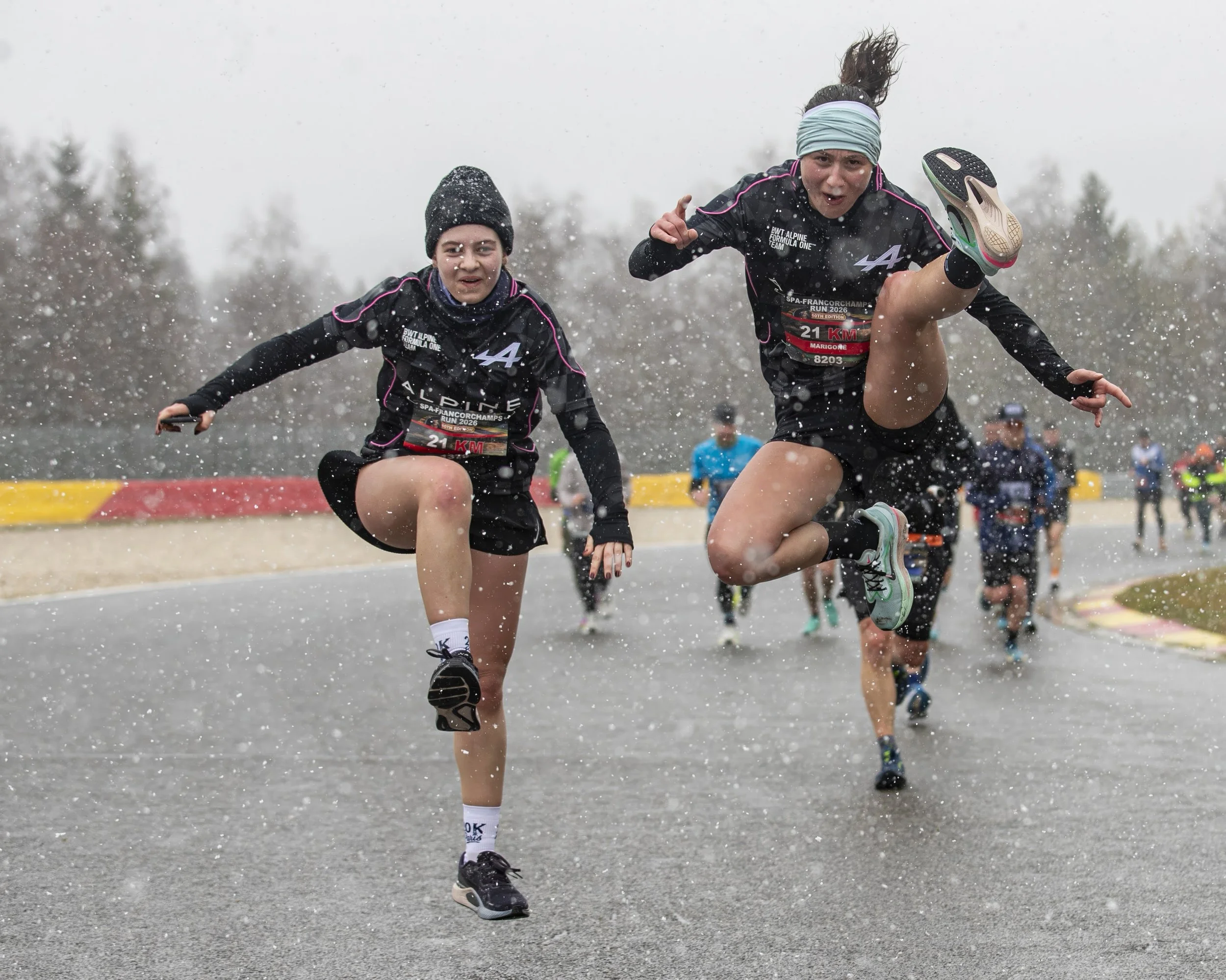 Two women running in a marathon through rain, wearing black athletic clothing, with trees in the background and rain droplets visible in the air.