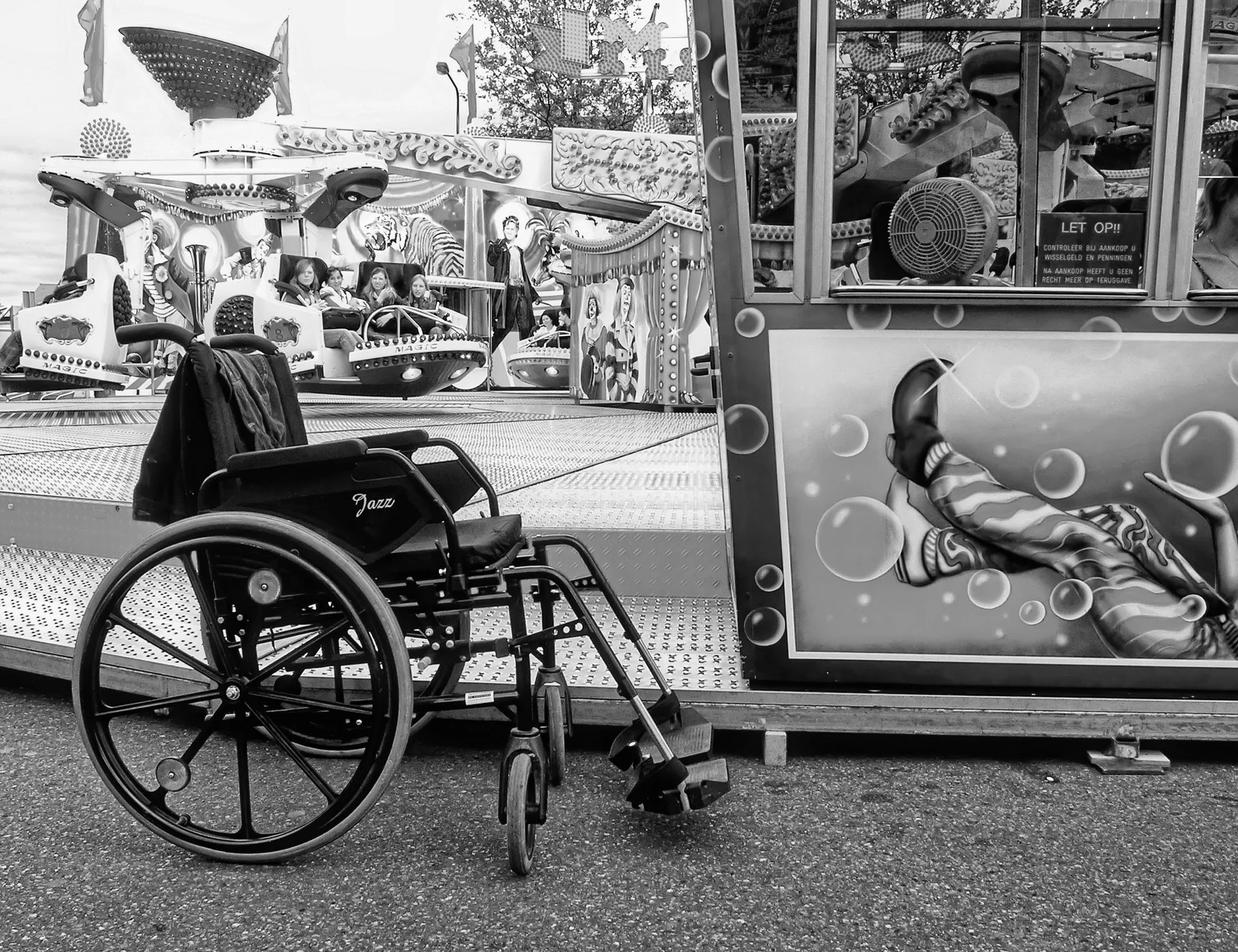 An amusement park ride with kid-sized cars and a person in the background. A disabled wheelchair is positioned in front of the ride. The ride features decorative bright lights and images of animals.