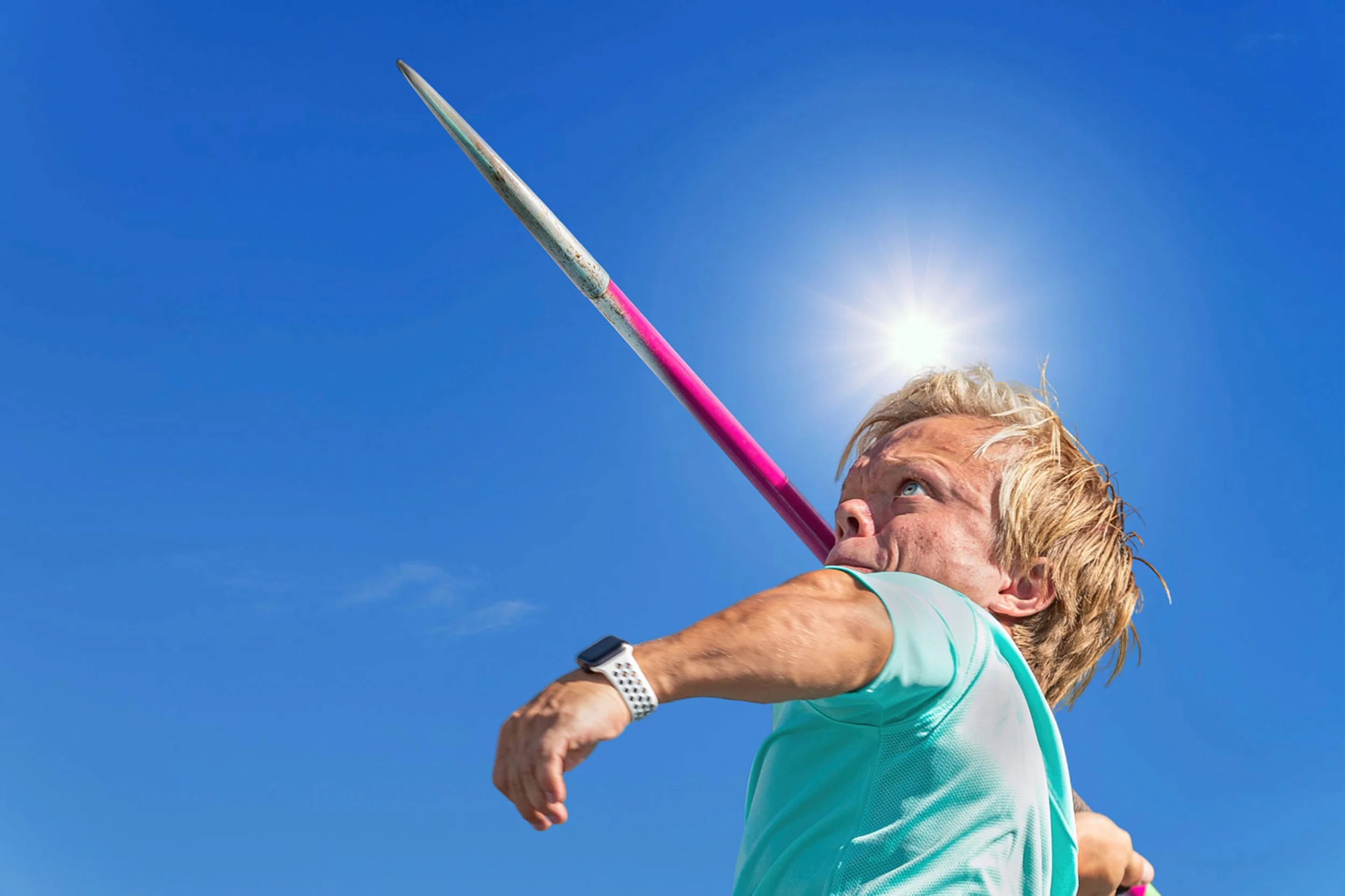 A middle-aged woman with blonde hair in a ponytail playing javelin outdoors against a bright blue sky. She is wearing a turquoise athletic shirt and a white wristwatch, and is in the motion of throwing the javelin upward.