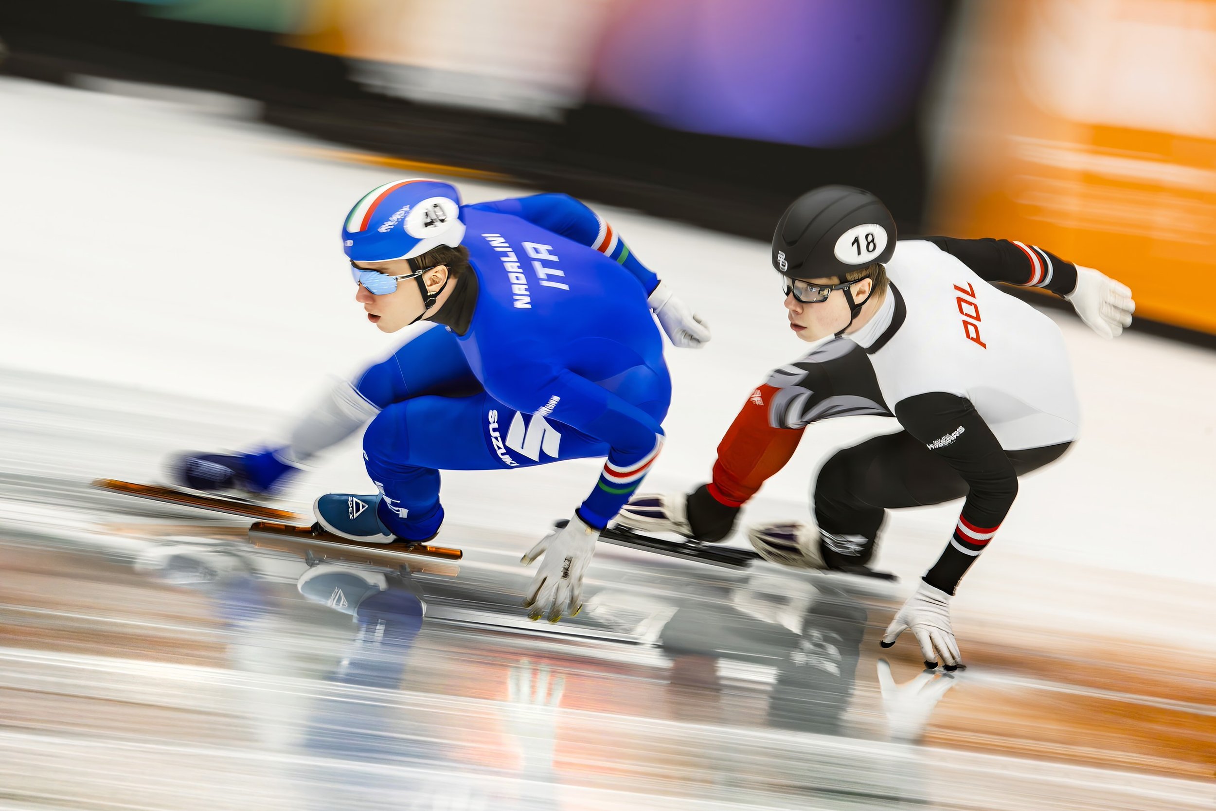 Two speed skaters in a race, leaning forward on the ice with motion blur in the background.