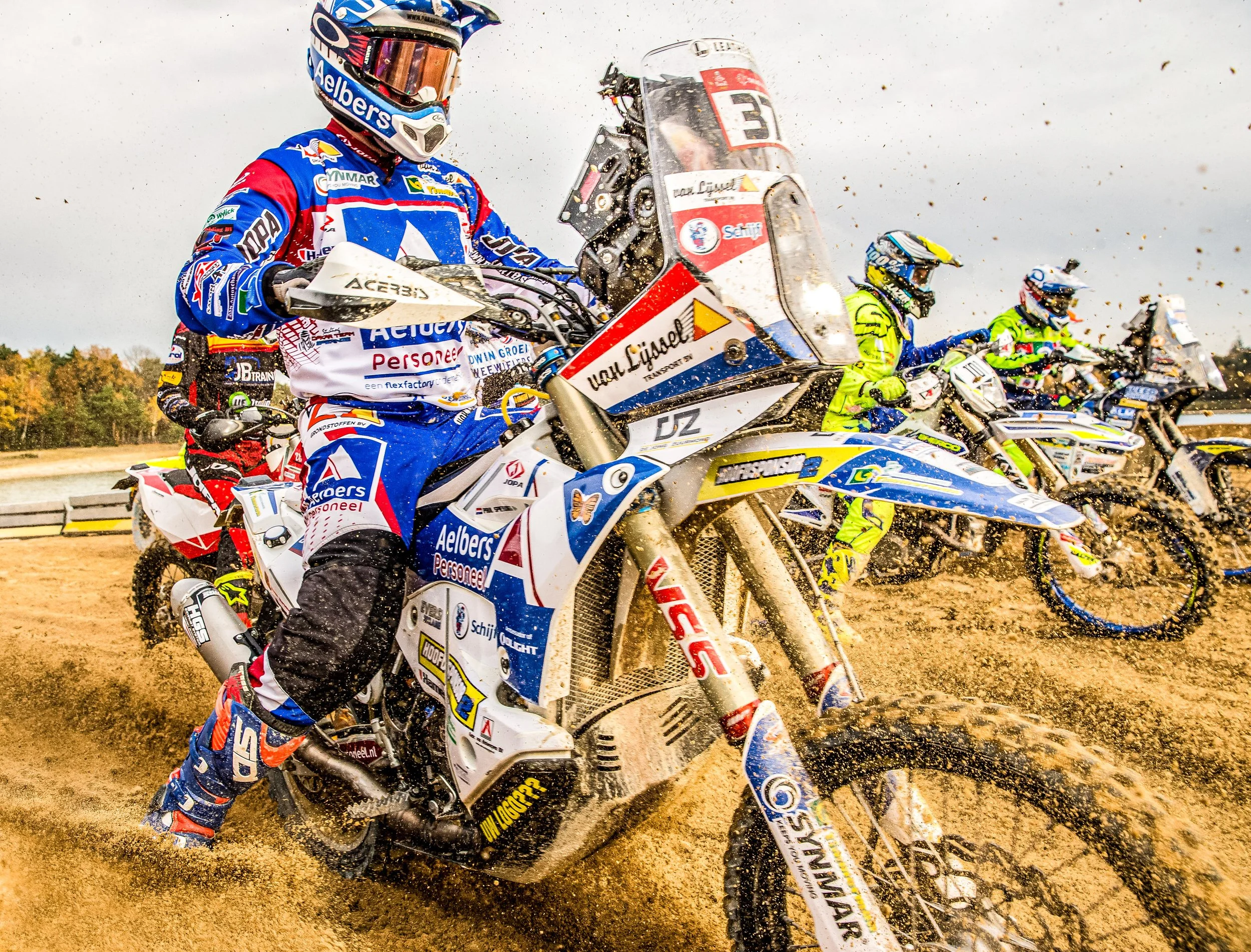 Motorcycle racer wearing a blue, white, and red suit and helmet, riding a dirt bike on a dirt track, kicking up dust and dirt particles during a race.