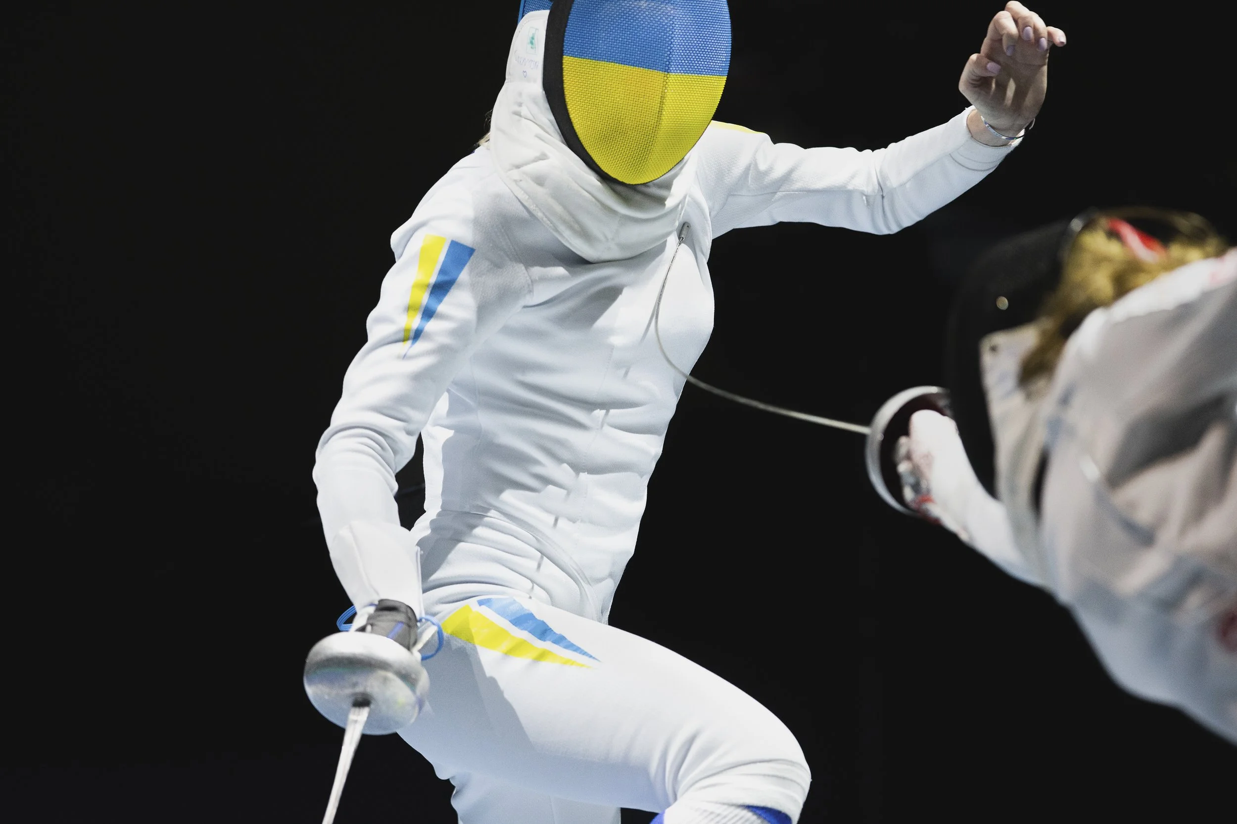A fencer dressed in white protective gear and a colorful mask is preparing to attack, holding a fencing sword, on a dark background.