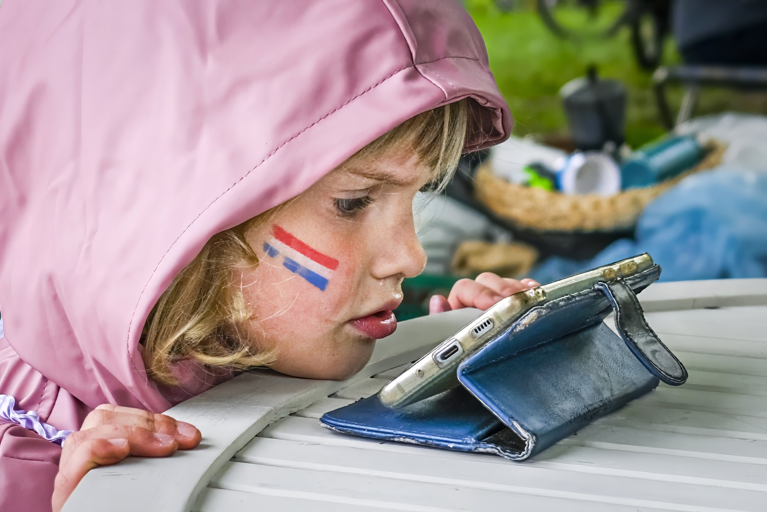 A young girl wearing a pink hooded jacket with a face painting of France's flag on her cheek, intently looking at her phone, resting her chin on a table, with a blurred outdoor setting in the background.