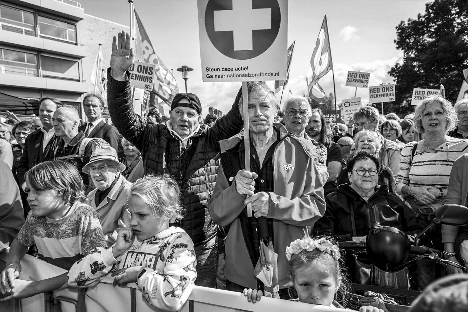 Crowd of people at a protest holding signs; man in the center holding a sign with a medical cross and Dutch text, raising his hand in a greeting gesture.