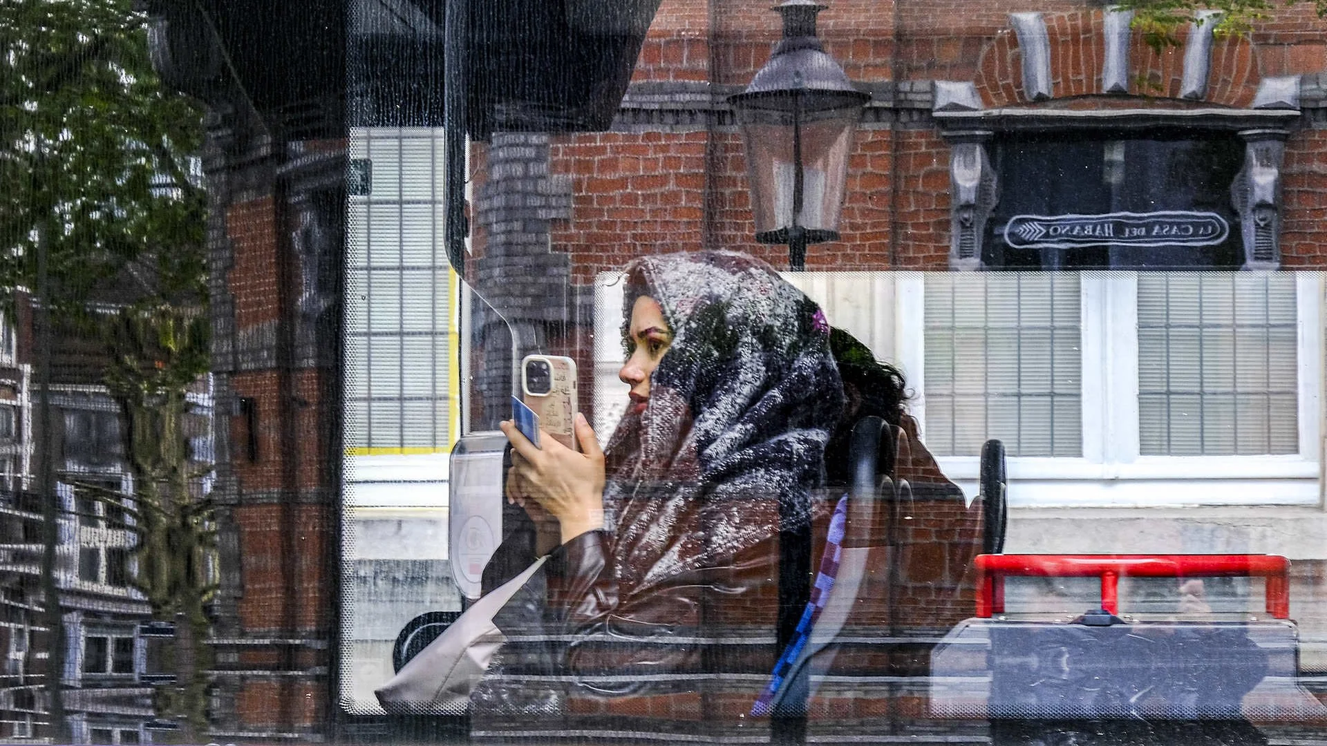 A woman with a patterned scarf over her head, sitting on a bus or train, taking a selfie with her phone, viewed through a rain-covered window with brick buildings and signage in the background.