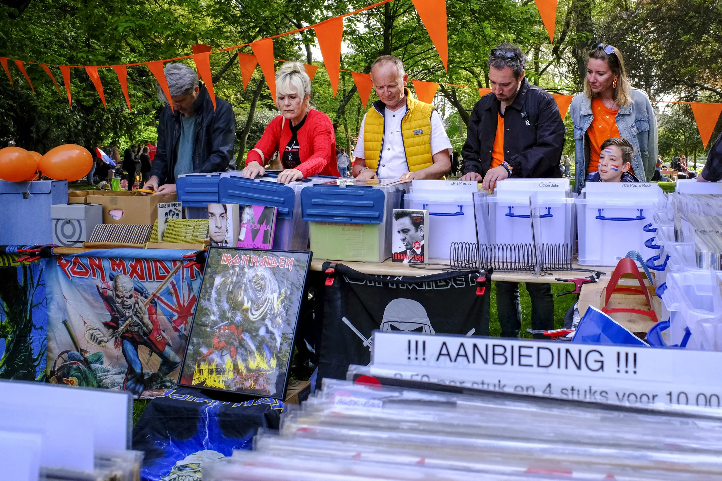 People browsing comic books and memorabilia at an outdoor market stall decorated with orange flags, in a park with green trees.