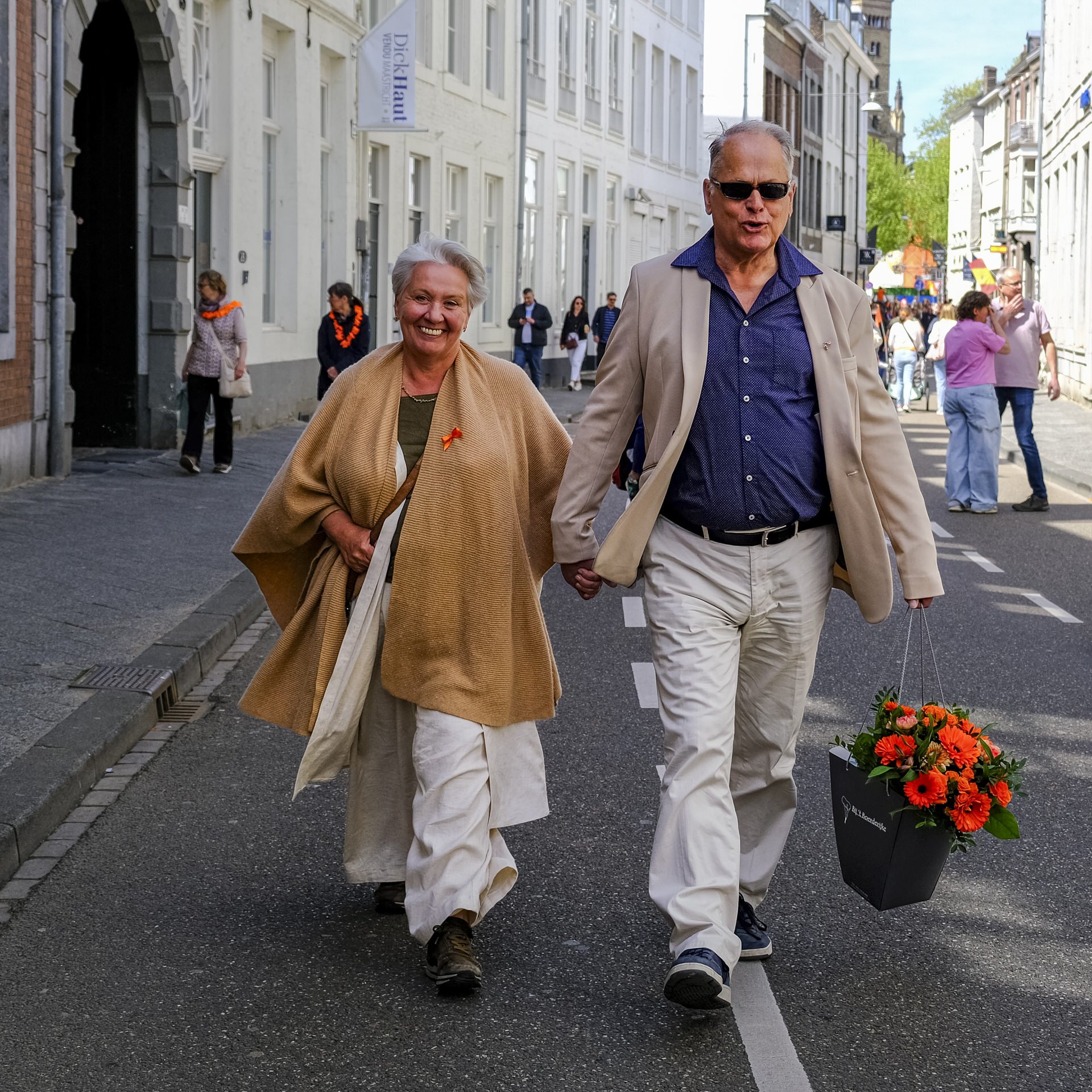 An elderly couple walking hand in hand down a city street during a parade or celebration, with store windows and people in the background. The woman is smiling and wearing a beige shawl with an orange ribbon, while the man has sunglasses and a beige 