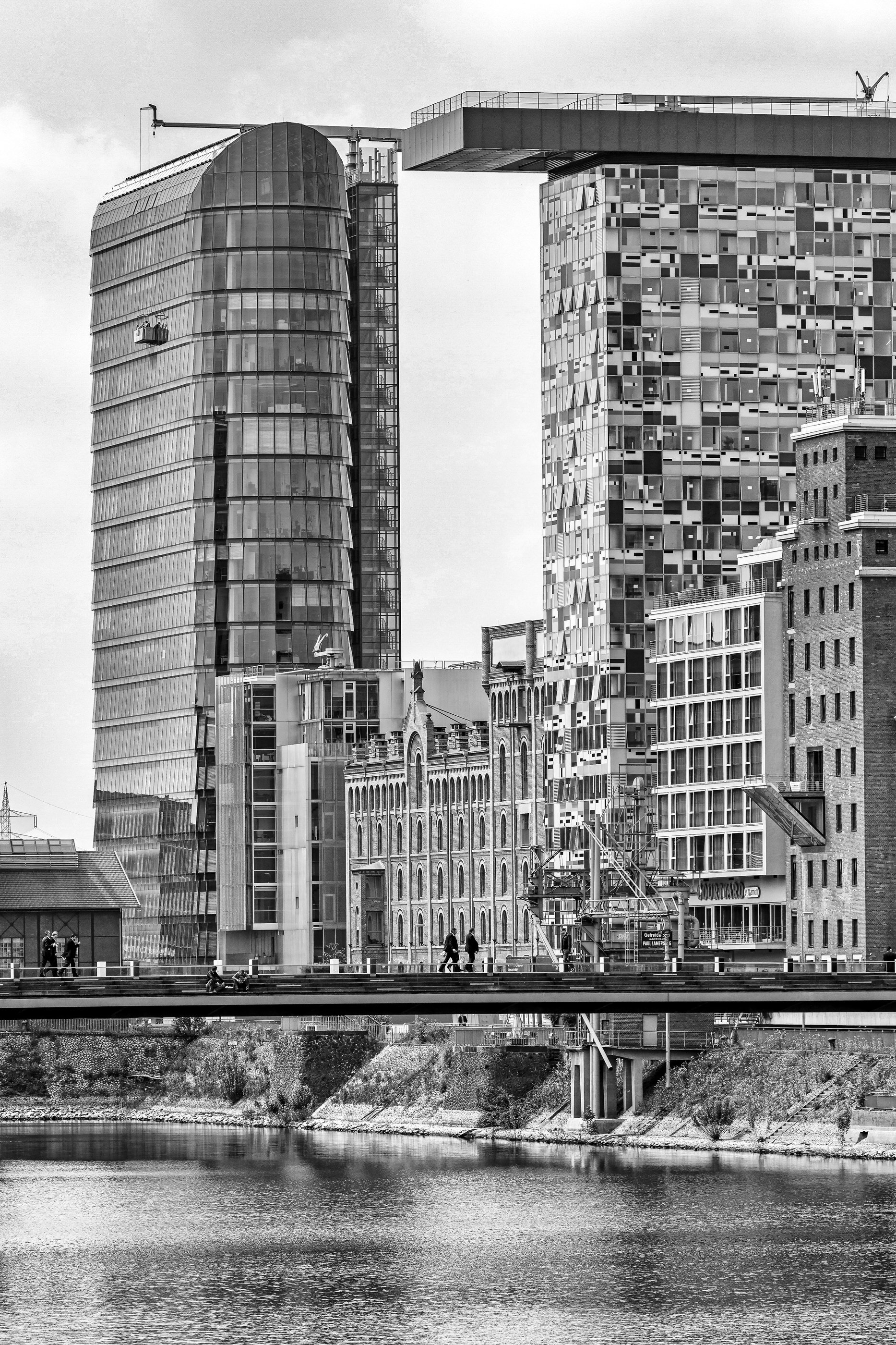 Black and white photo of modern and historic buildings along a river in an urban city, with people walking on a bridge over the river.