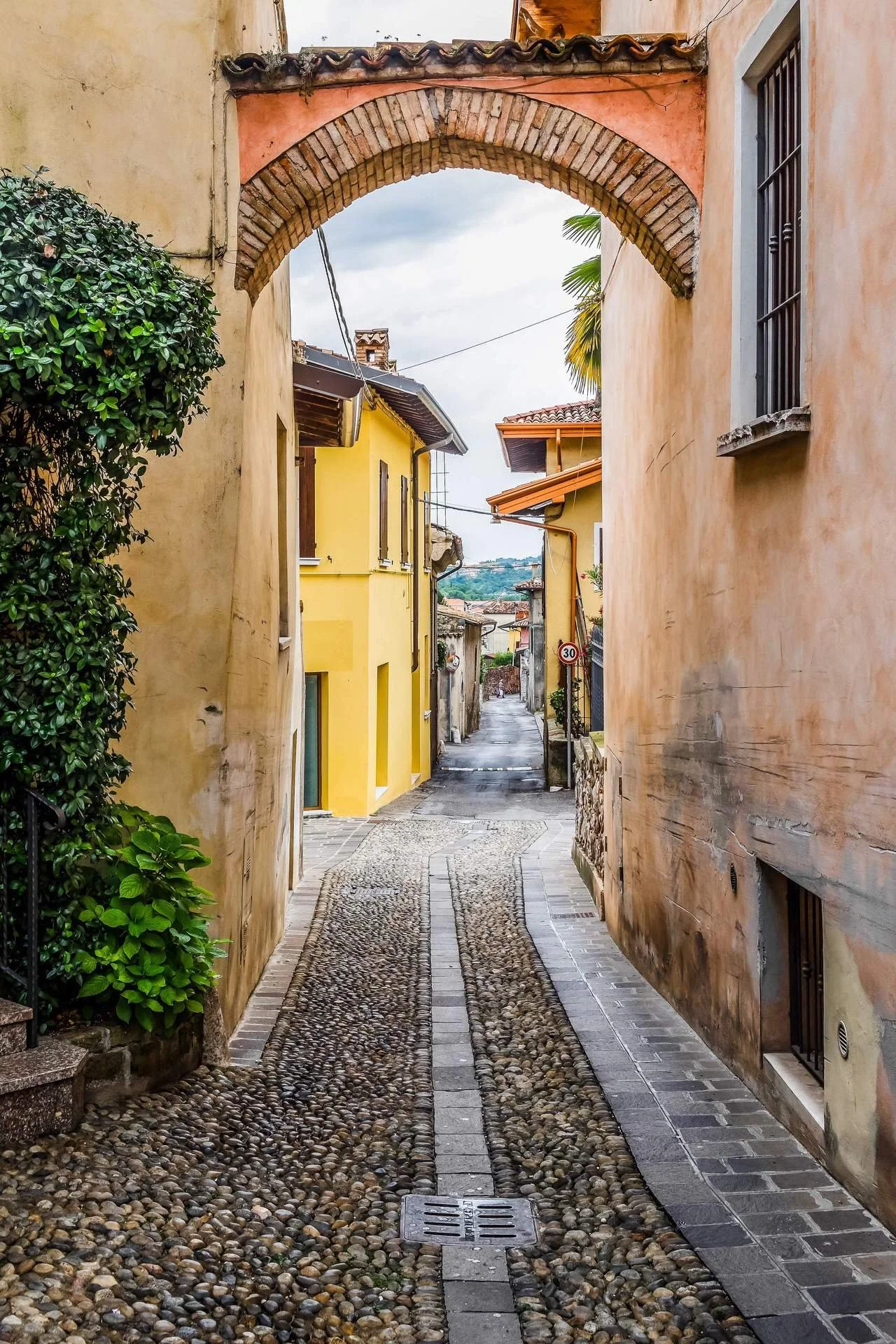 A narrow cobblestone street in a European village, lined with colorful buildings and an archway overhead.