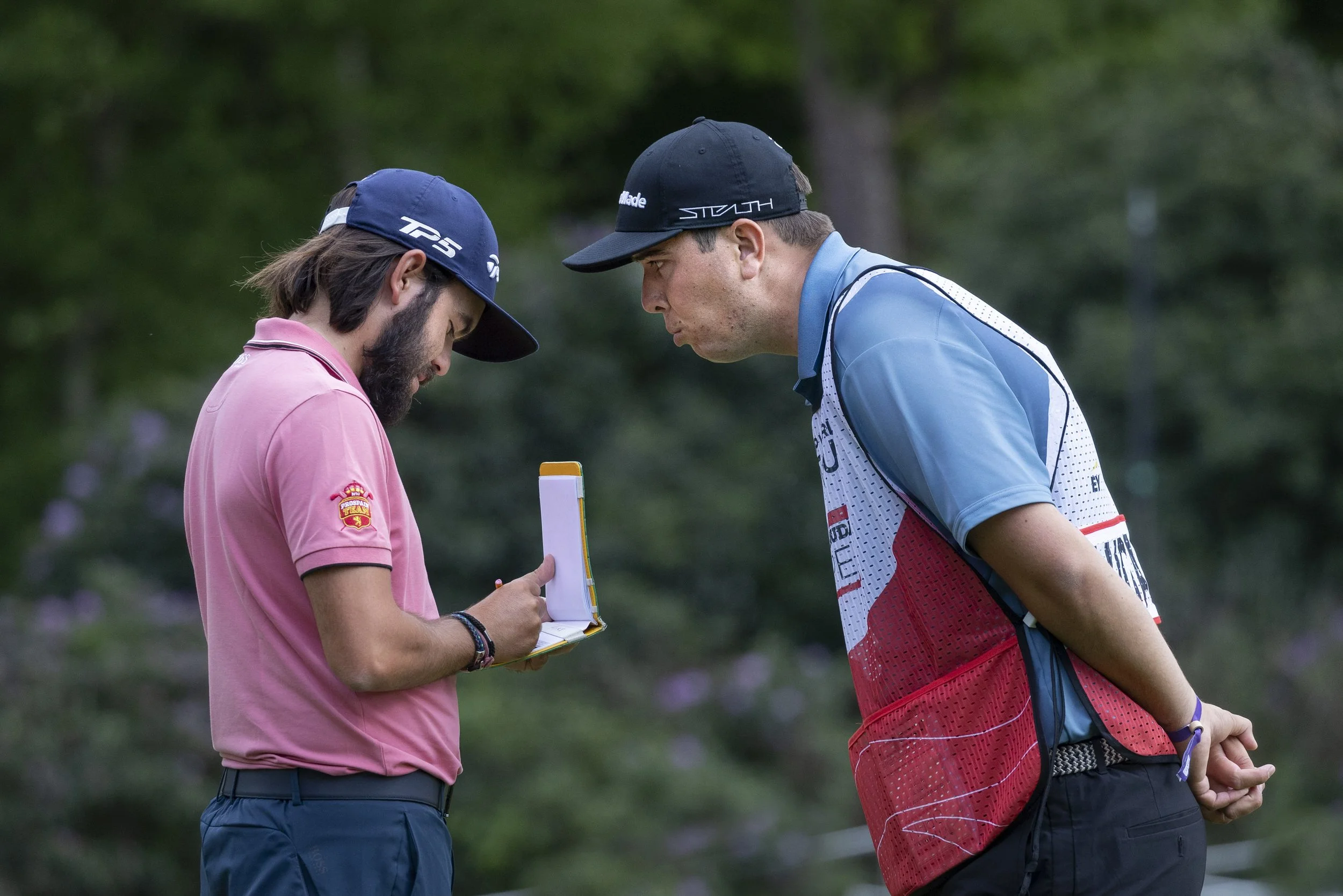 Two male golfers are engaged in a discussion during a golf game. The golfer on the left wears a pink shirt and a blue cap, holding a scorecard, while the golfer on the right wears a blue and red shirt with a black cap. They are outdoors with a blurre