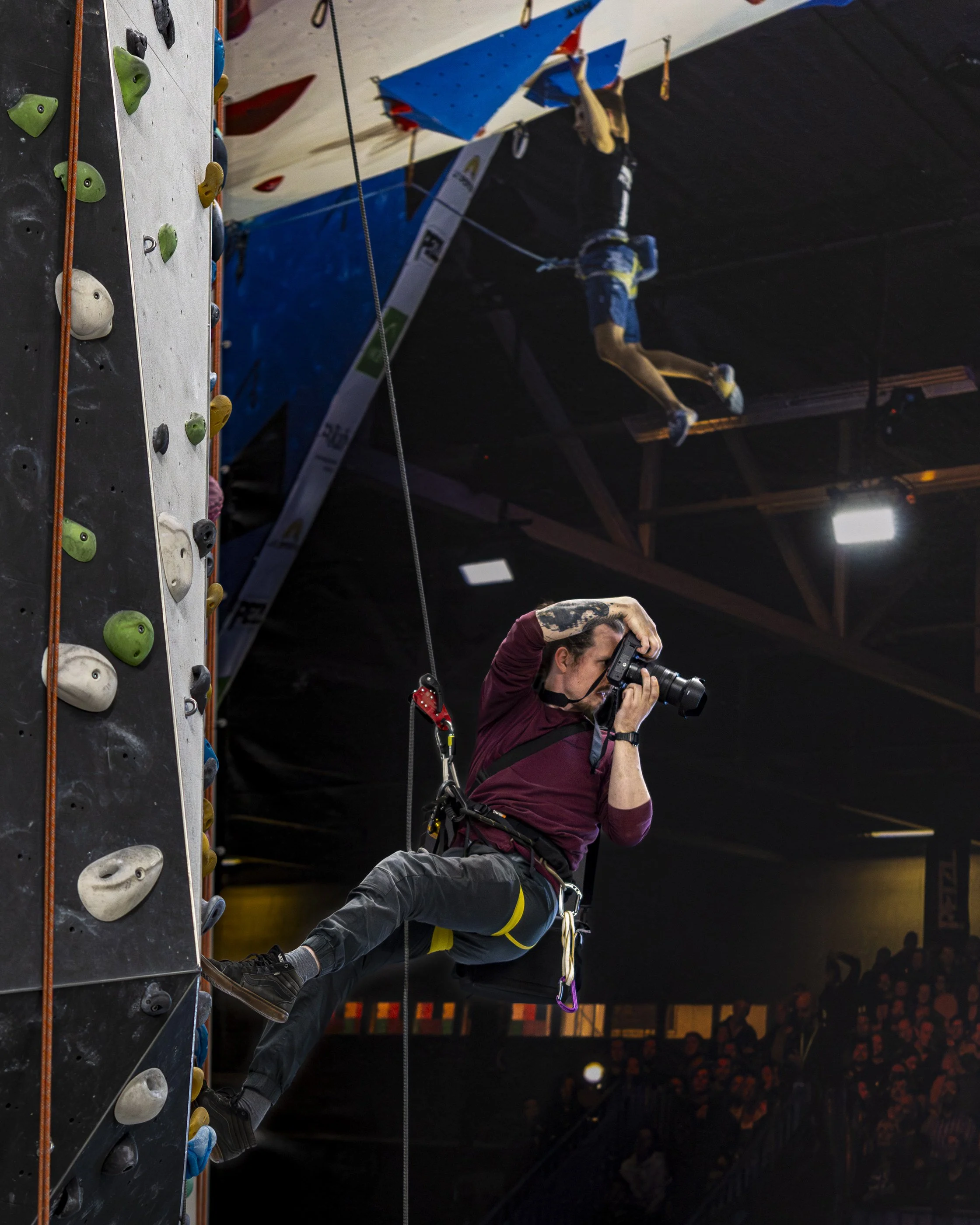 An indoor rock climbing gym with climbers. One person is climbing on the wall with colorful holds, and a photographer is taking pictures of the climbers. The climbers are secured with harnesses and ropes.