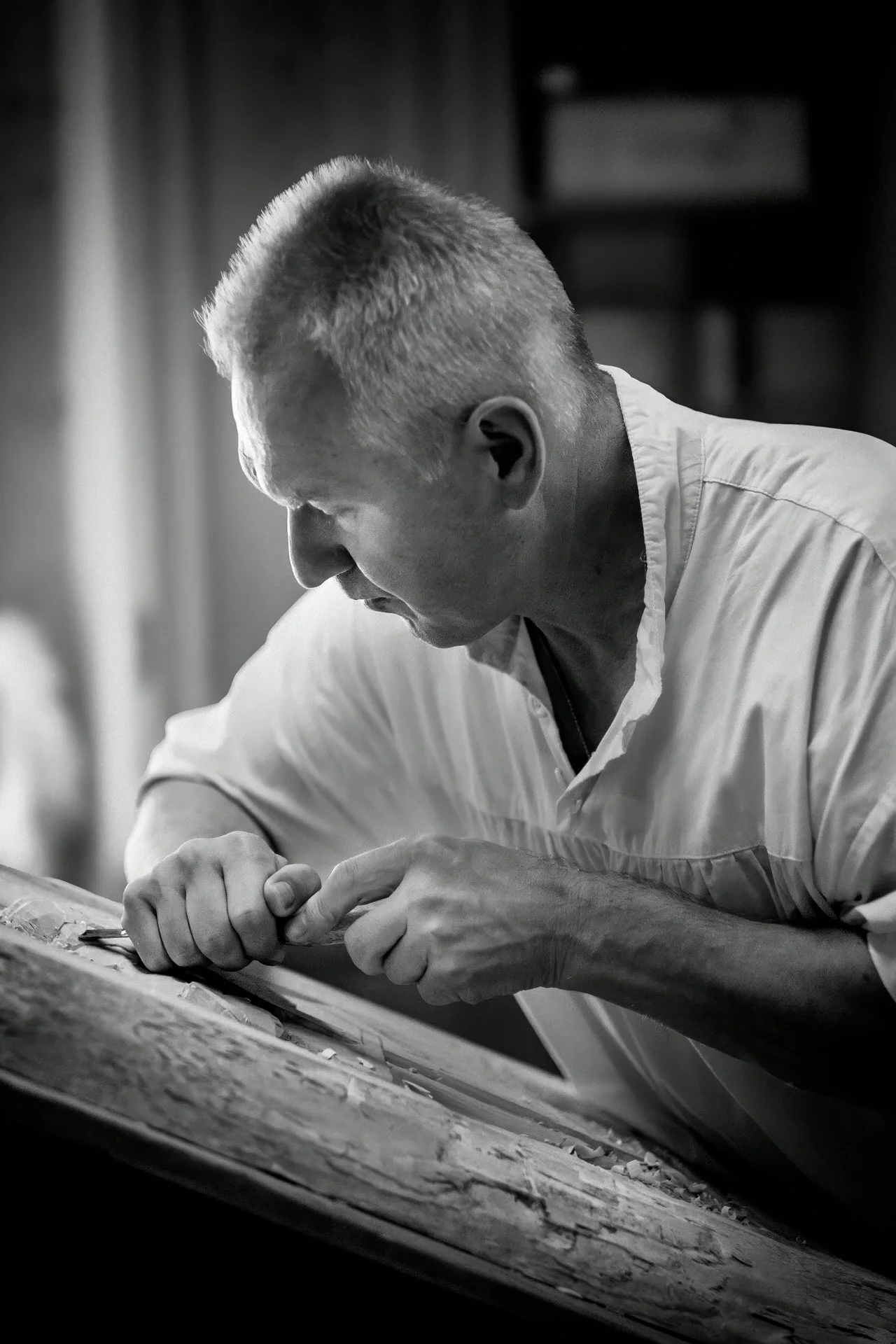 A man with short hair and a white shirt is working on a piece of art or craft, using his hands to shape or manipulate material on a flat surface. The background is slightly blurred.
