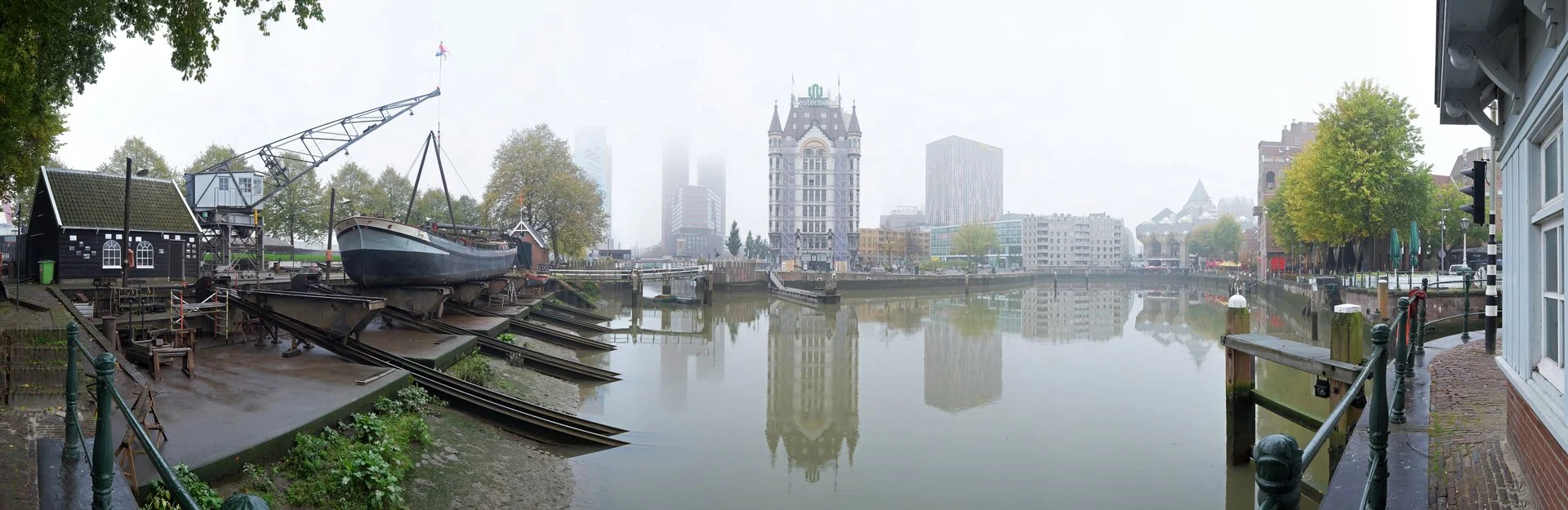 A cityscape view along a waterway with buildings, trees, and foggy sky in the background, seen from a riverside walkway with railing and brick buildings.