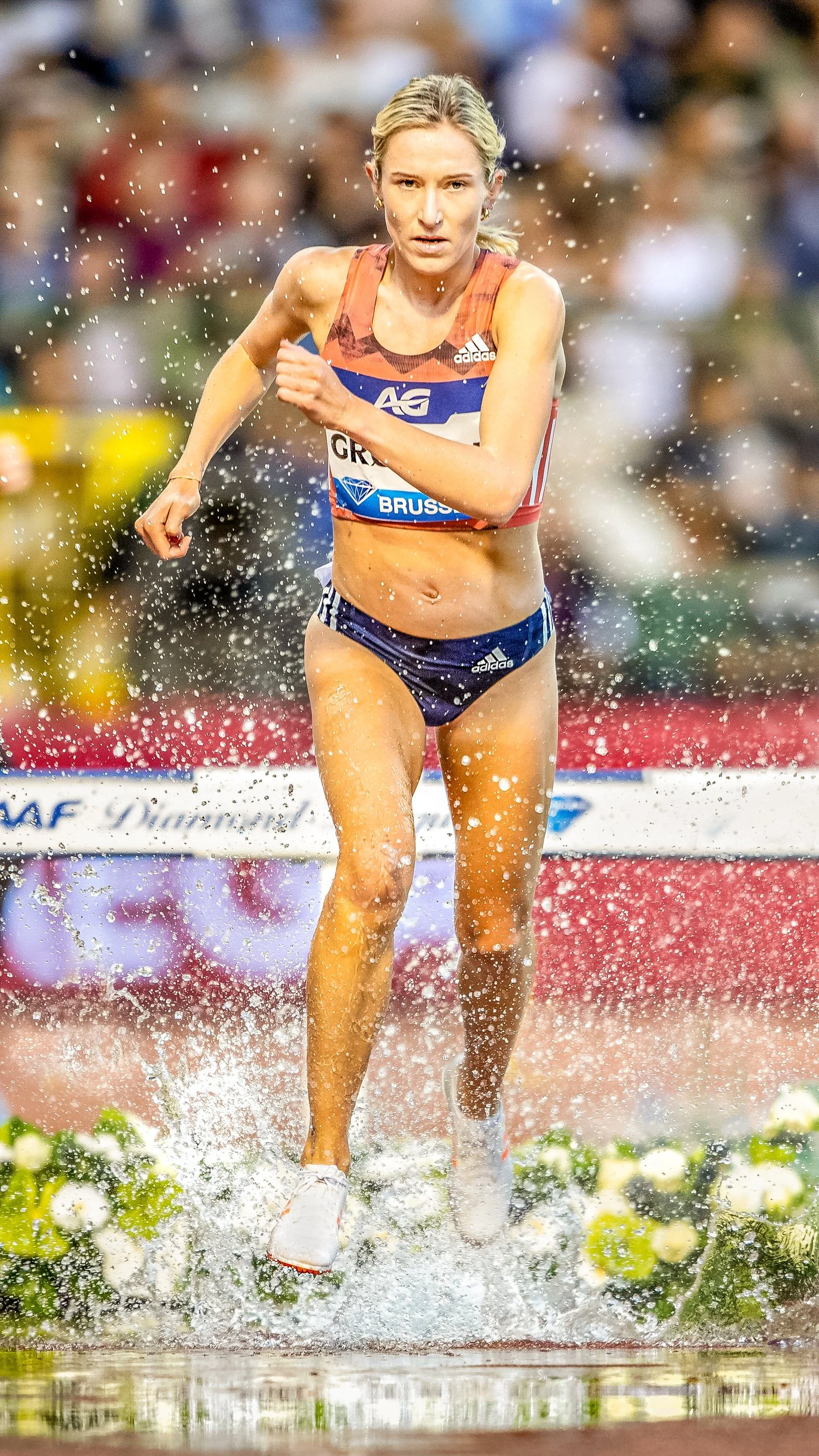 Female athlete running through water during a track race, with a blurred crowd of spectators in the background.