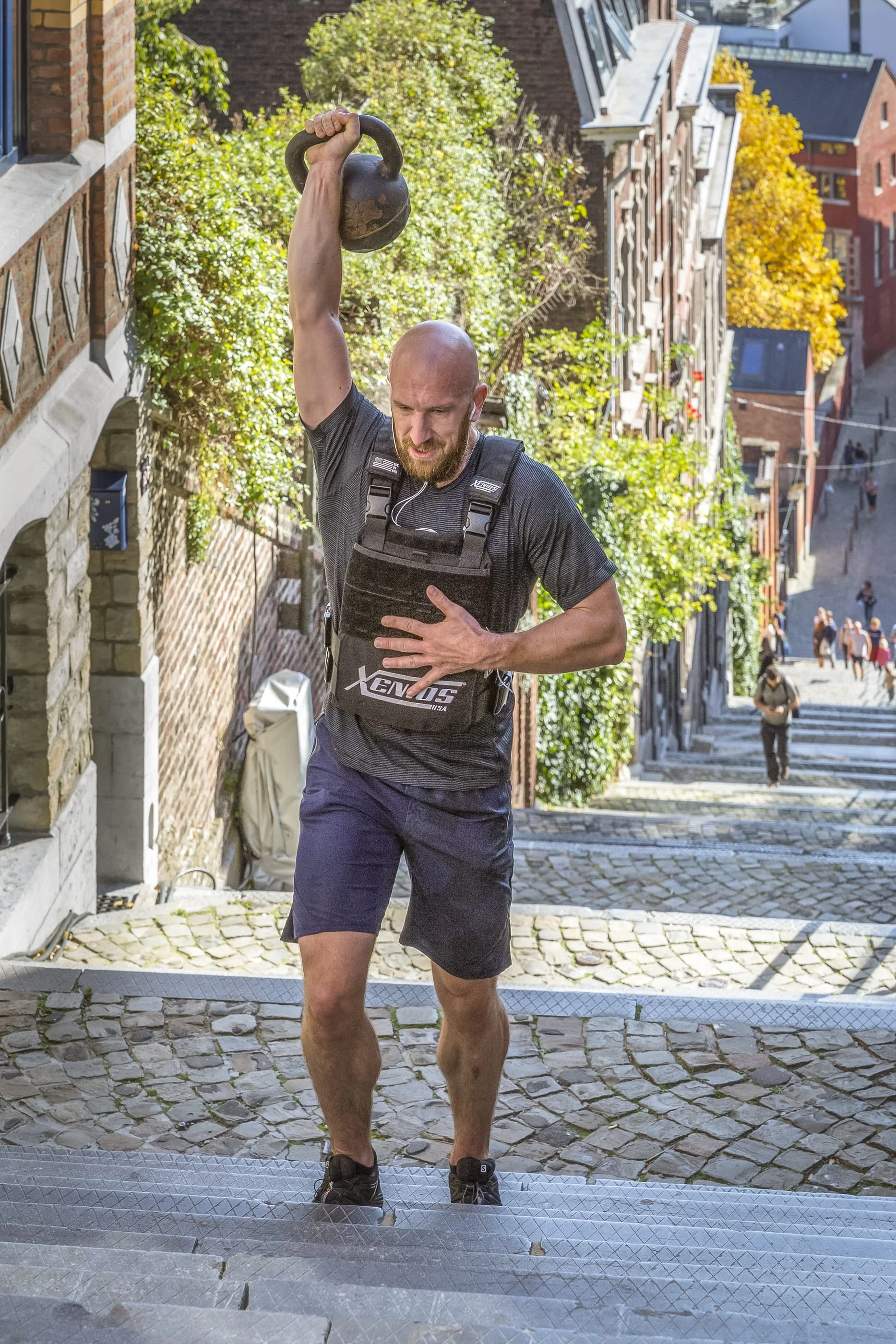 Man running up cobblestone stairs outdoors, carrying a kettlebell overhead with one hand, wearing a black athletic shirt with a chest pack, black shorts, and headphones.
