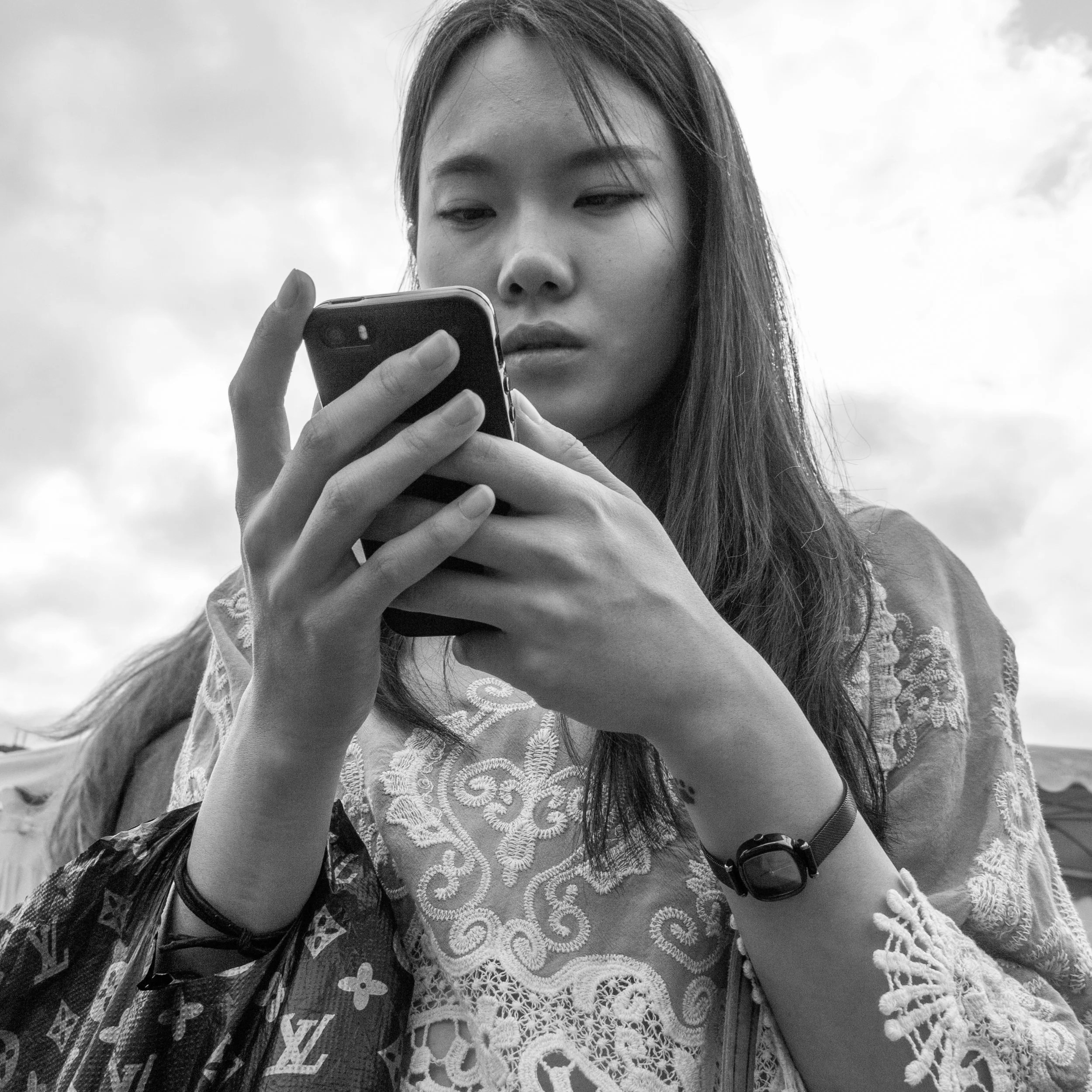 A woman with long hair looking at her phone outdoors during daytime, wearing a lace top and accessories, with a cloudy sky in the background.