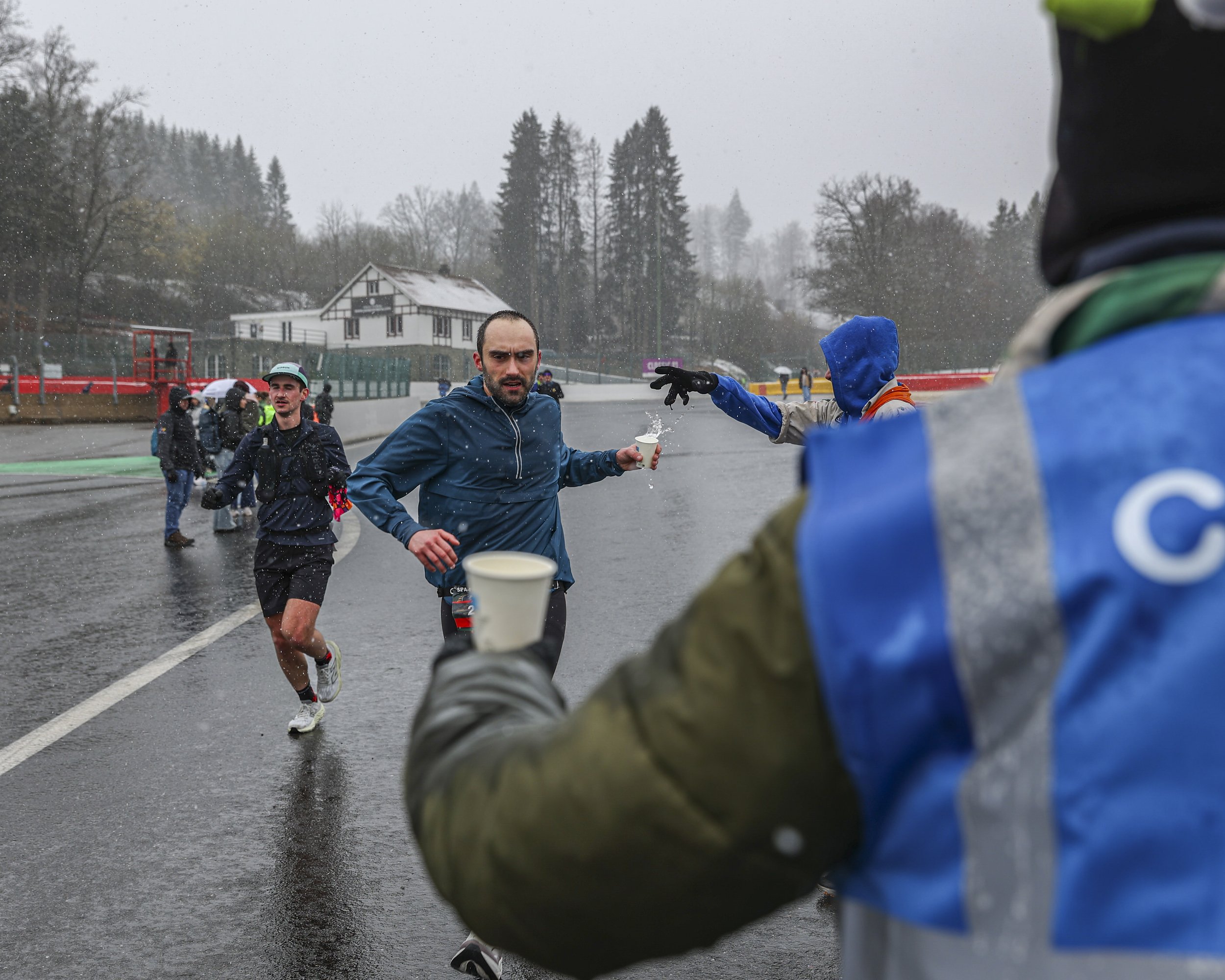 A group of marathon runners, including a man in a blue jacket, running on a wet road during rainy weather with trees and a house in the background.