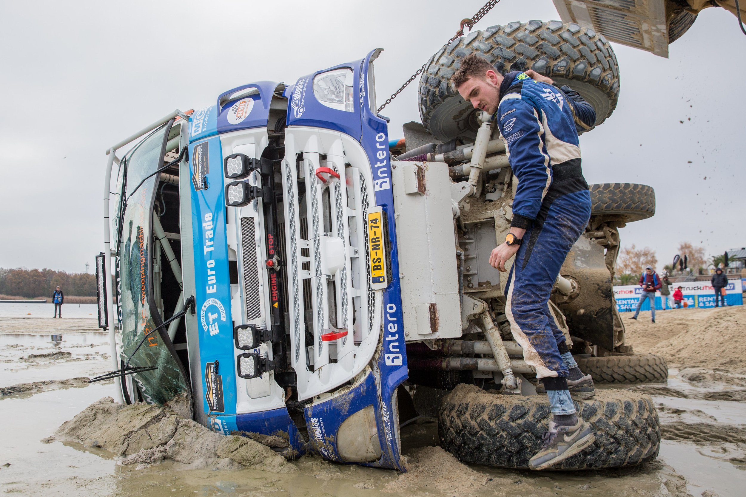 A race truck is overturned on muddy terrain with muddy water and dirt splattered around. A man in blue racing gear is inspecting or repairing the truck, which is resting on its side.