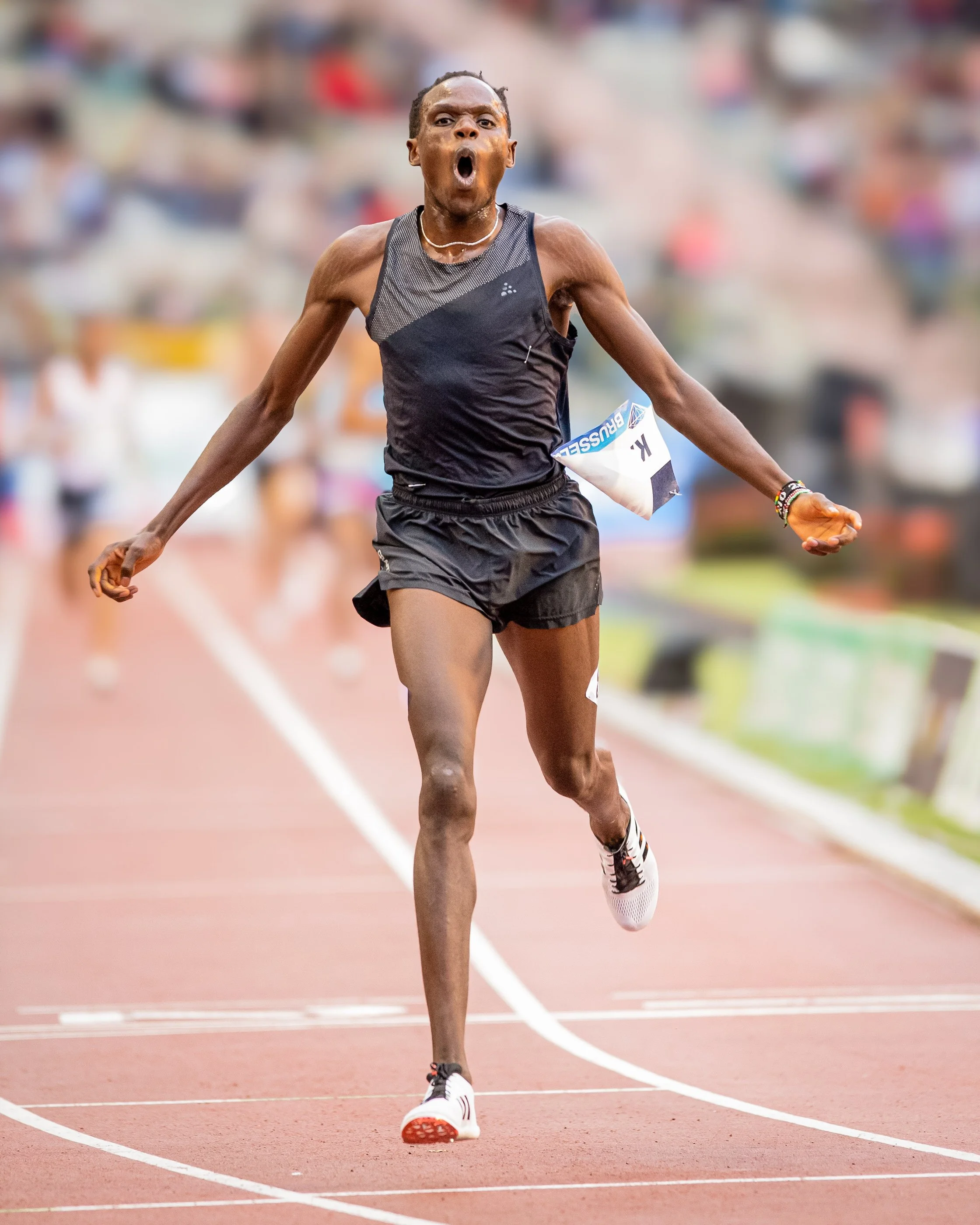 A male athlete racing on a track with a crowd in the background, showing a determined and intense expression.