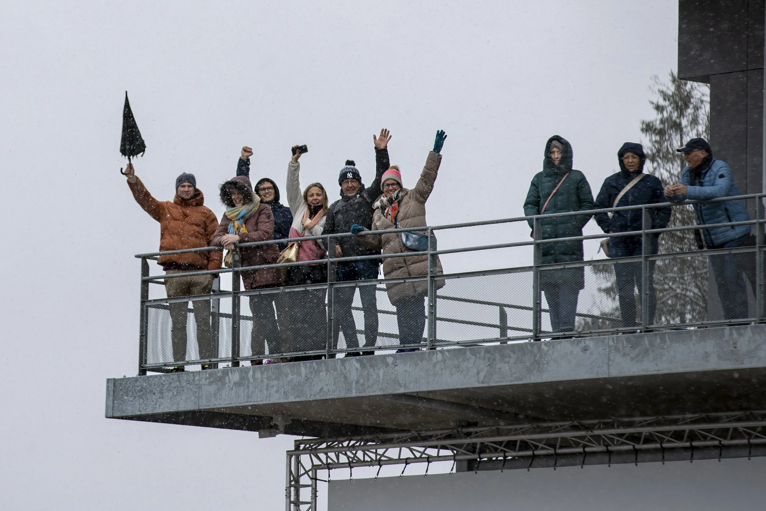 A group of eight people standing on an outdoor balcony in cold weather, wearing winter clothing, some with hoods or hats, celebrating or enjoying the snow. Snowfall is visible in the air.