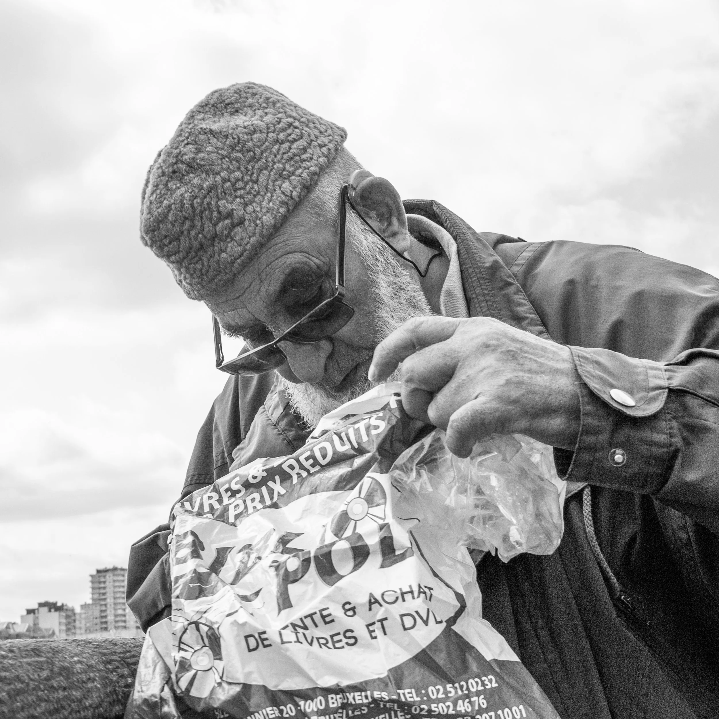 A black and white photo of an elderly man with glasses, wearing a warm hat with ear flaps and a jacket, holding a crumpled bag, with city buildings in the background.
