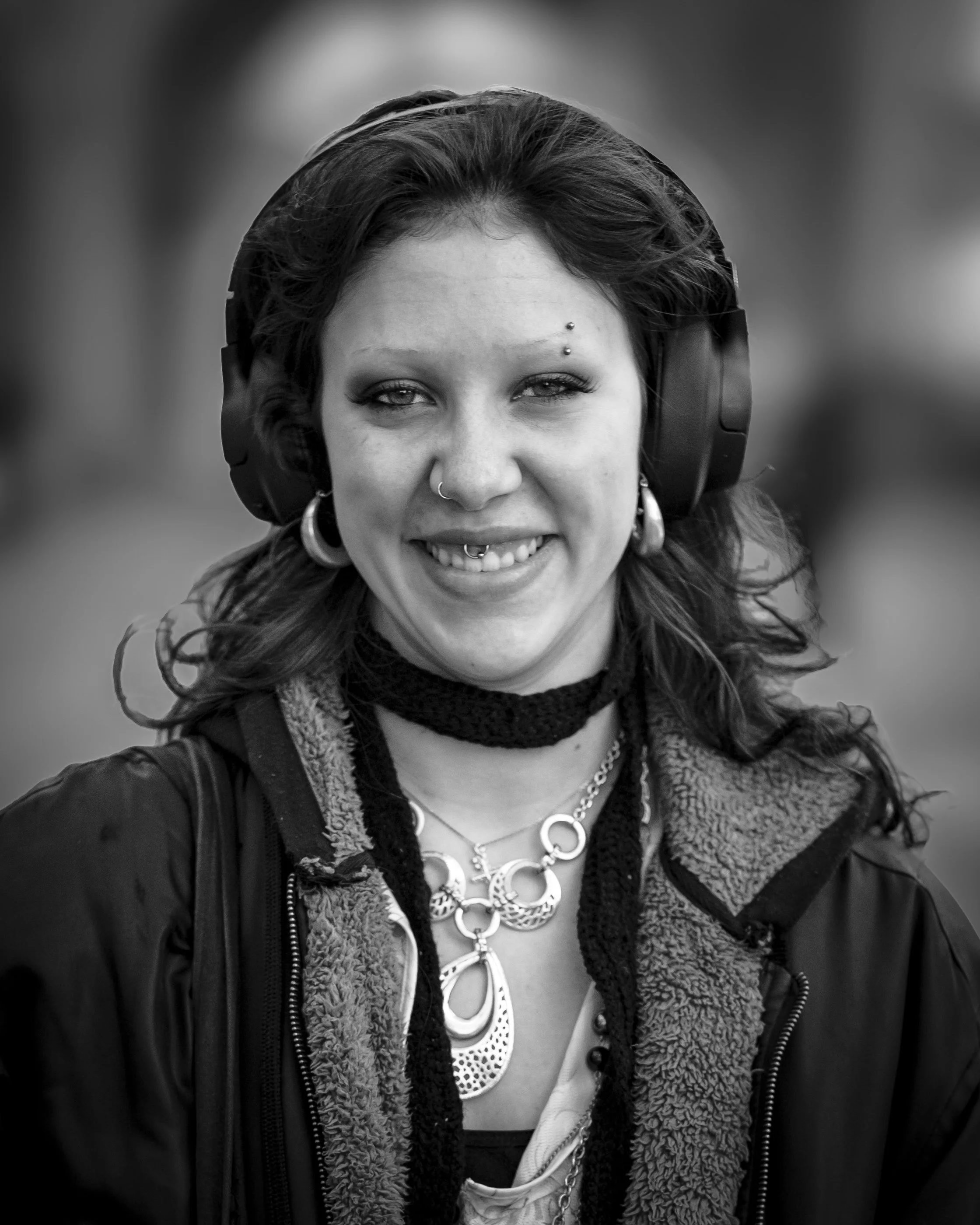 A woman with dark hair, wearing large headphones, multiple piercings including a nose ring and eyebrow piercings, layered necklaces, a scarf, and a jacket with a fleece-lined collar.