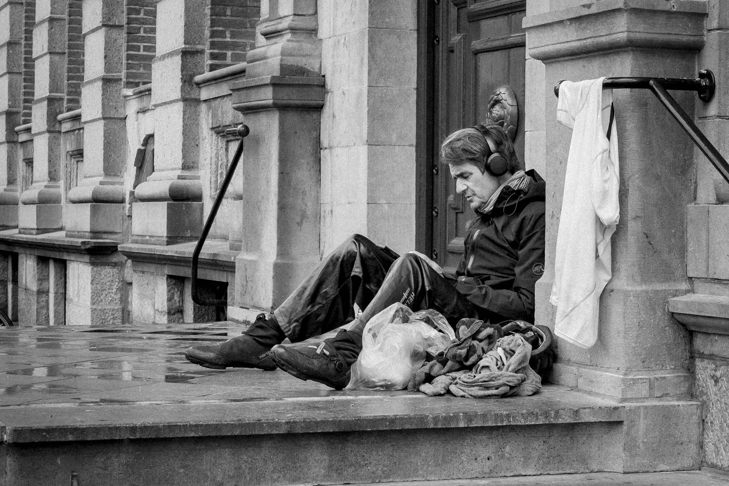 A man sitting on steps outside a building, wearing headphones and reading a book with a bag and clothes beside him. The building has ornate stone and brick architecture.