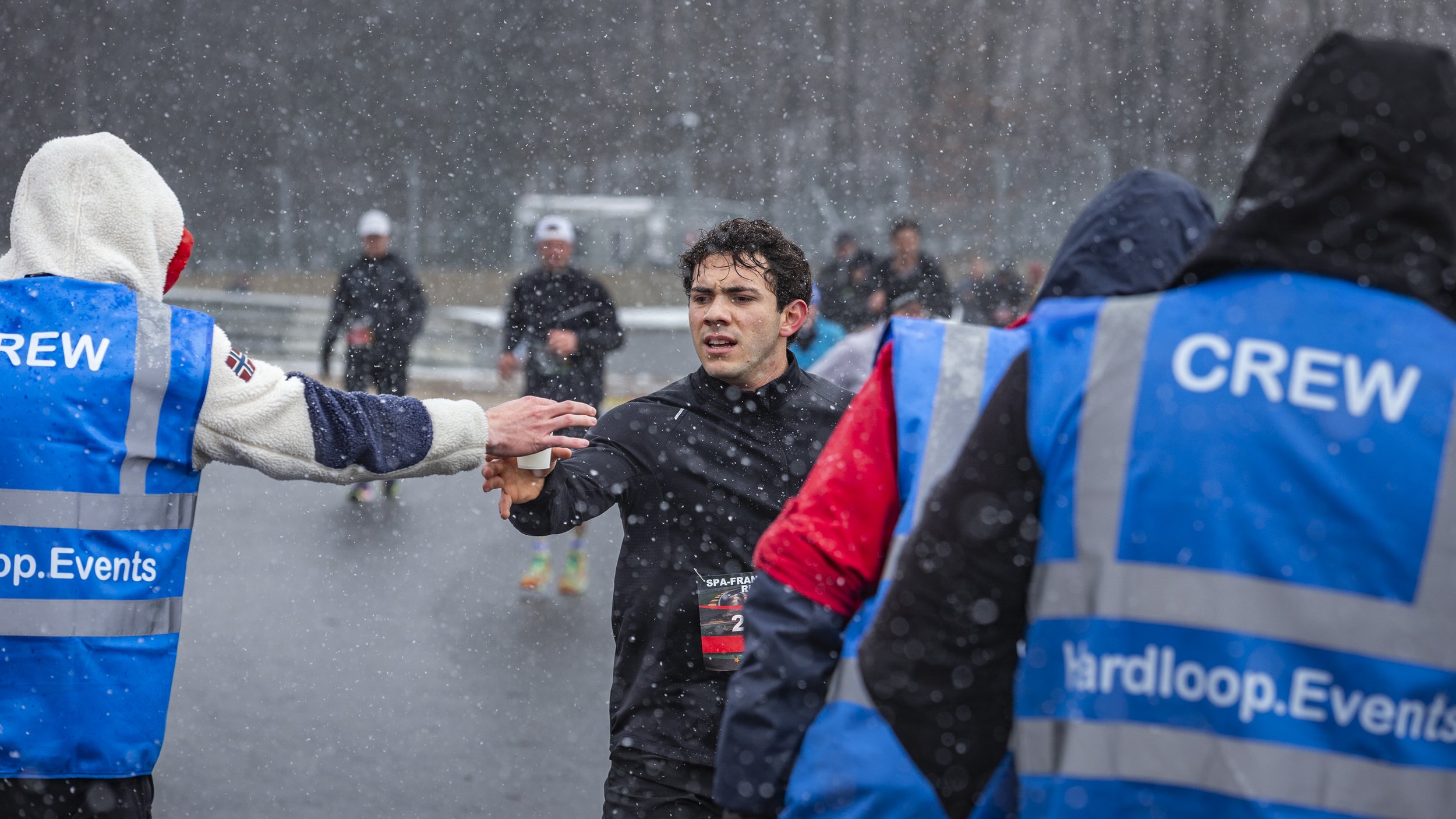 A marathon runner reaches out to accept water from race volunteers on a rainy day, with other runners in the background.