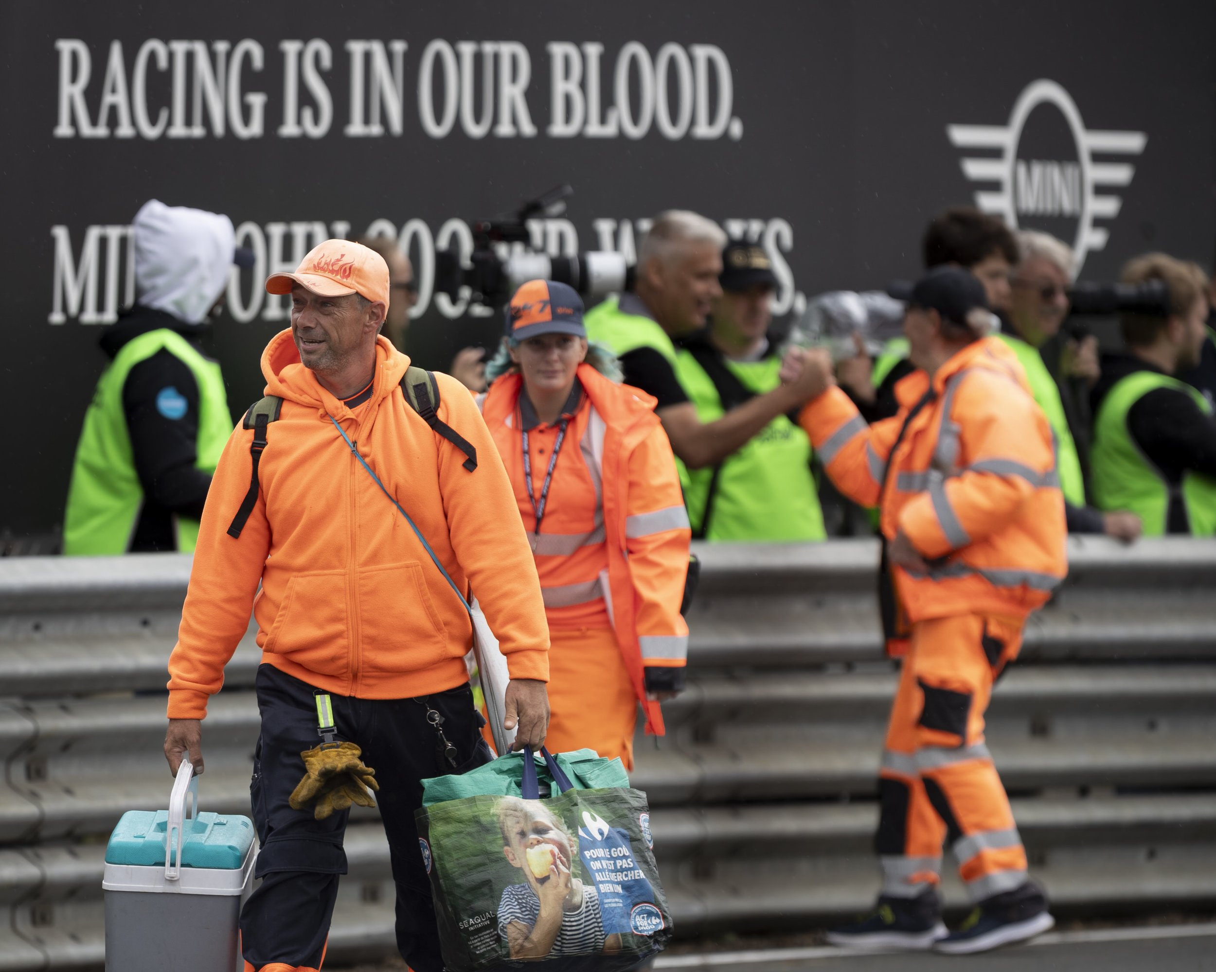 Group of people dressed in bright orange and neon green safety vests and jackets walking past a large black wall with white text and a logo. Some are carrying bags and cooling boxes, with a man in the foreground wearing an orange hoodie and carrying 