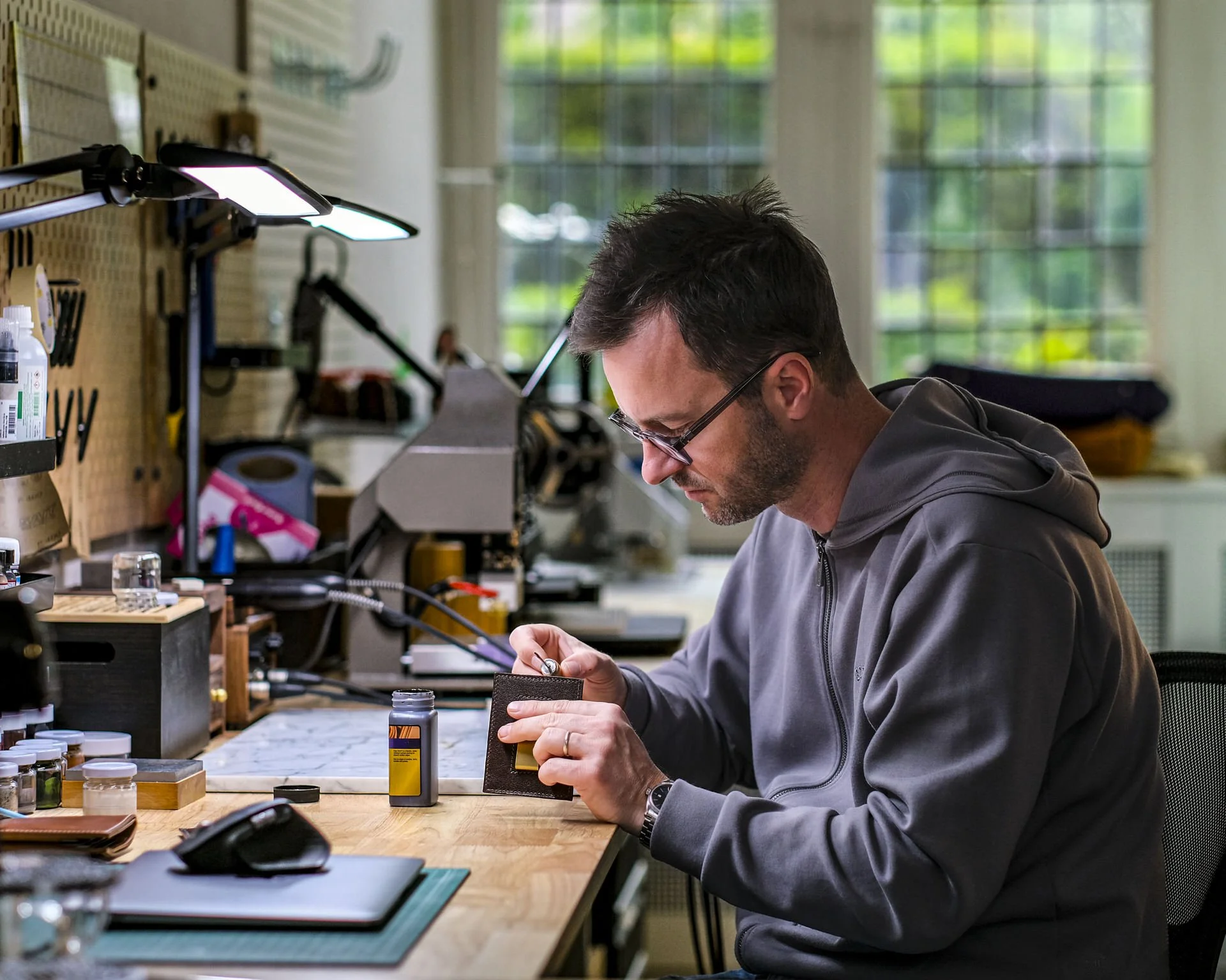 Man working on a leather wallet at a worktable in a workshop, surrounded by tools, bottles, and supplies for leather crafting, with a pegboard and large windows in the background.