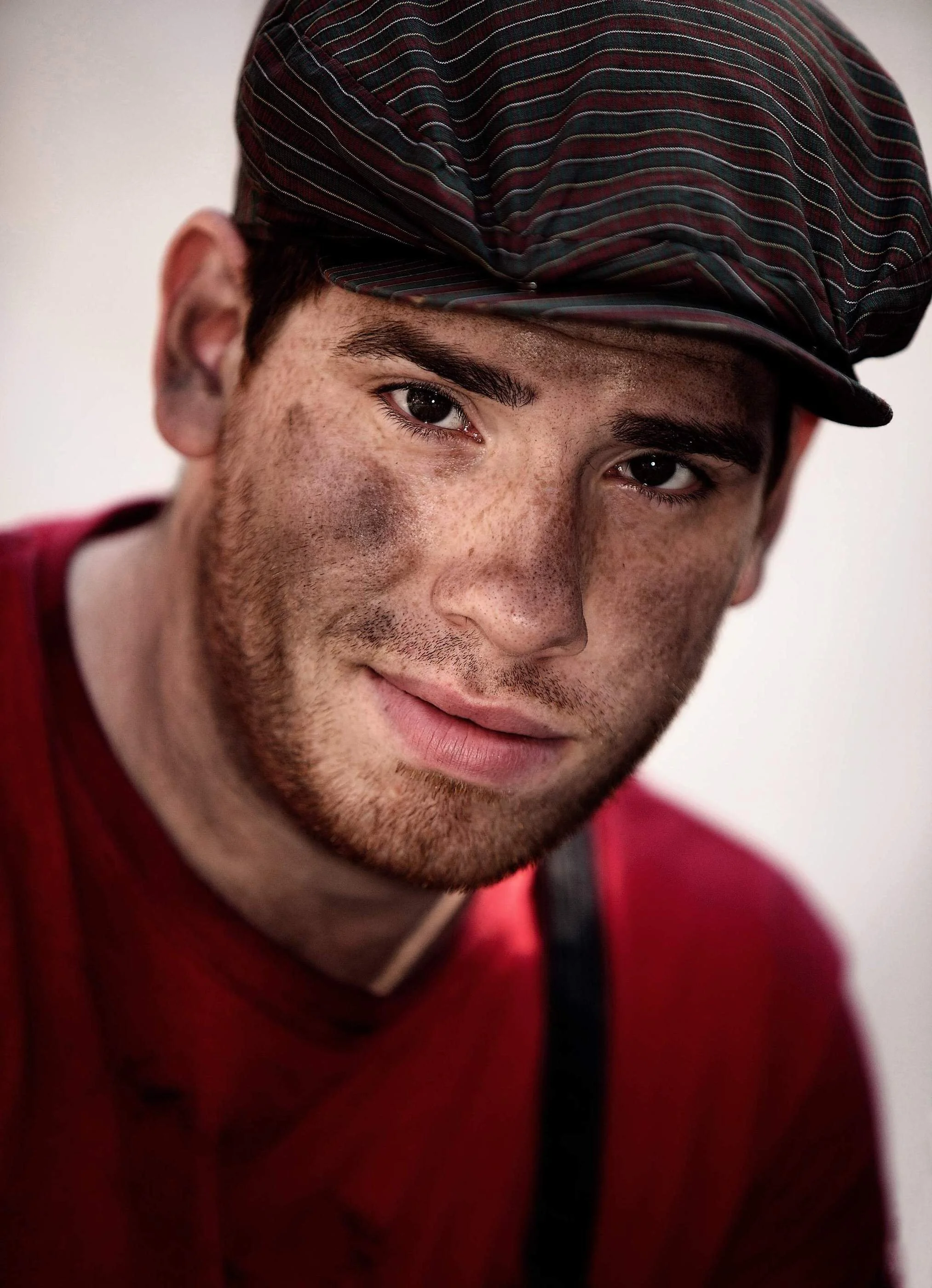 Close-up of a young man with dark hair, freckles, and dark eyes wearing a striped cap and a red shirt.