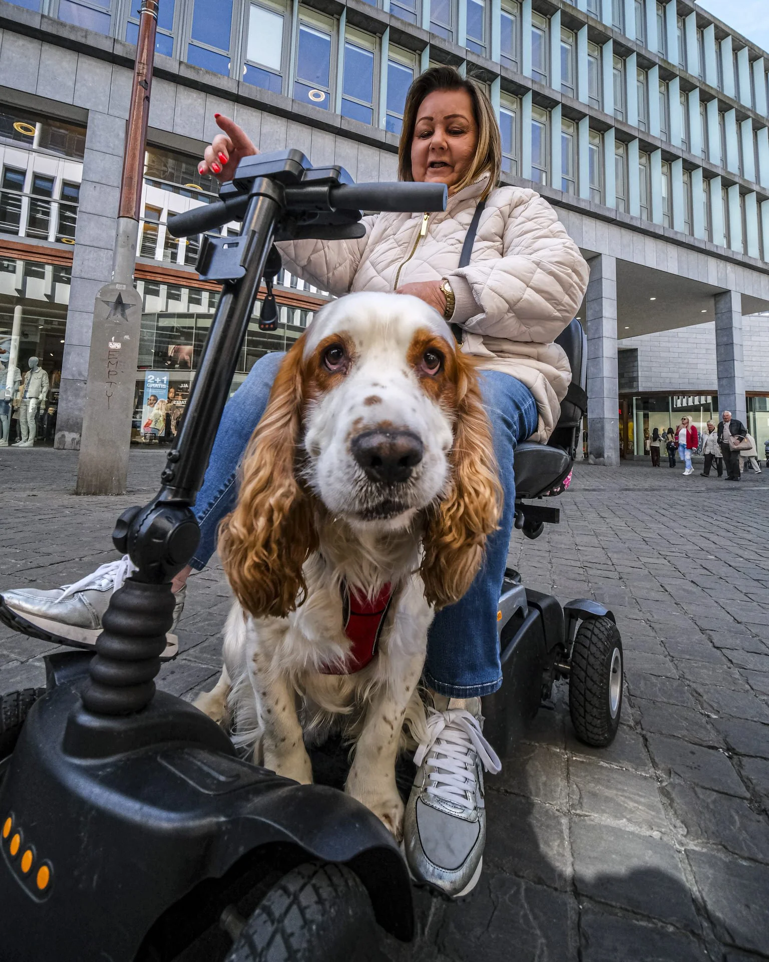A woman with short hair and a beige jacket seated on a mobility scooter in a city plaza, with a large dog in front of her, and people walking in the background.